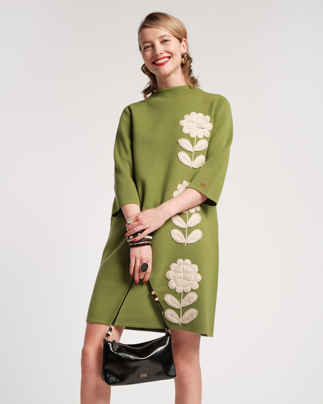 A person with shoulder-length hair stands with their back to the camera, wearing a green Frances Valentine Annie Dress Posy Embroidery Merino Wool Fern. The knee-length dress features three-quarter sleeves and is paired with black flats. Several bracelets adorn the person’s wrist, and they are positioned against a plain white background.