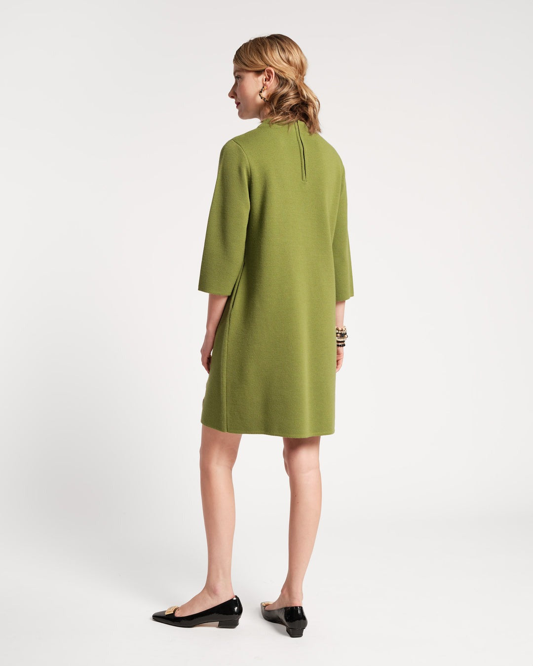 A person with shoulder-length hair stands with their back to the camera, wearing a green Frances Valentine Annie Dress Posy Embroidery Merino Wool Fern. The knee-length dress features three-quarter sleeves and is paired with black flats. Several bracelets adorn the person’s wrist, and they are positioned against a plain white background.