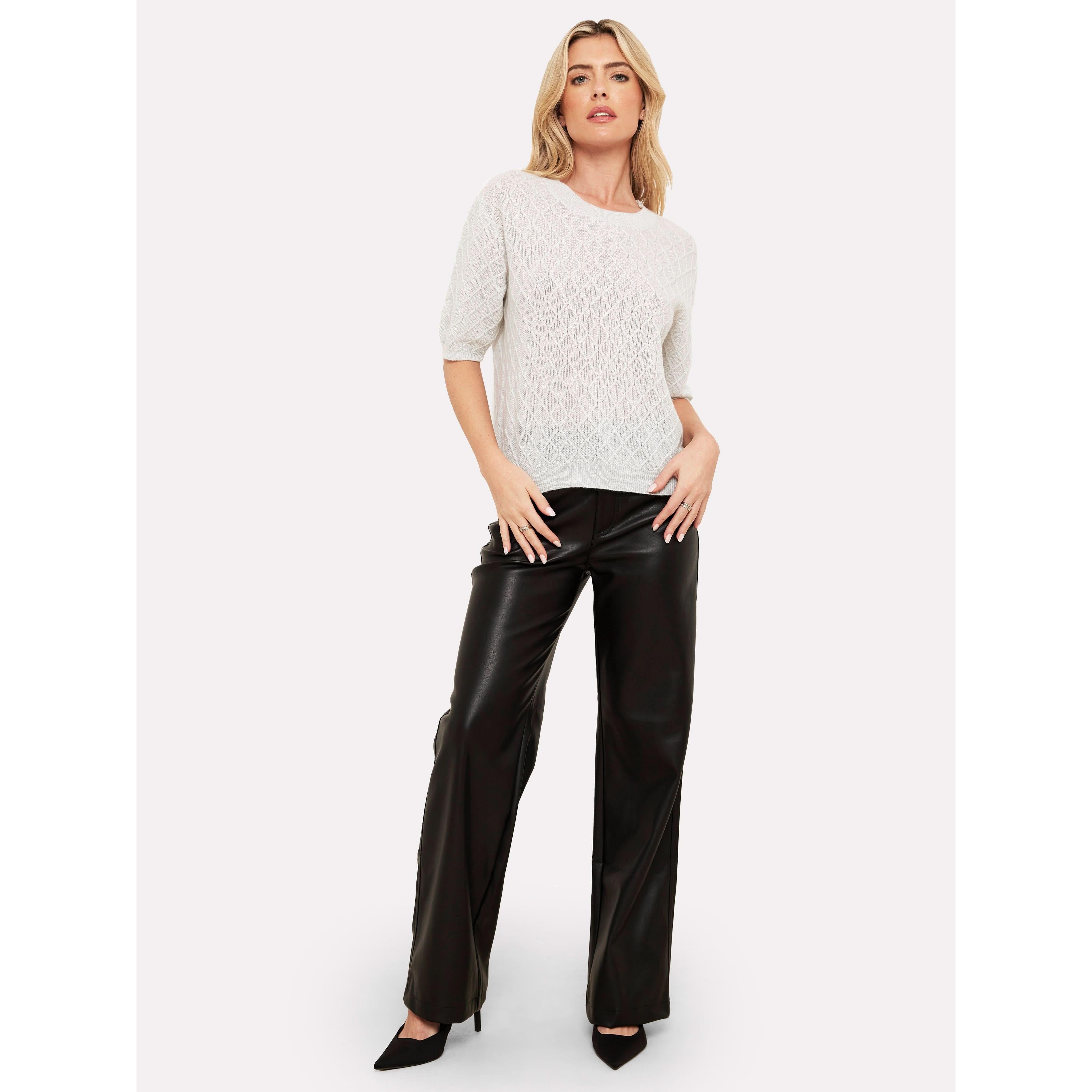 A person dressed in the Brodie Cashmere Cable Pearl Tee and black leather pants stands against a plain white background. With one hand in their pocket and wearing black heels, they effortlessly exude elegance.