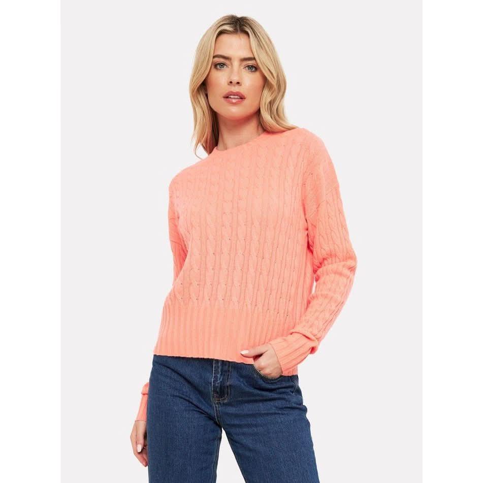 A person wearing the Brodie Cashmere Cathy Cashmere Cable Knit sweater in neon coral, featuring a ribbed crew neckline, stands with one hand in their pocket against a plain white background. They have long, wavy blonde hair and are paired with dark blue jeans.