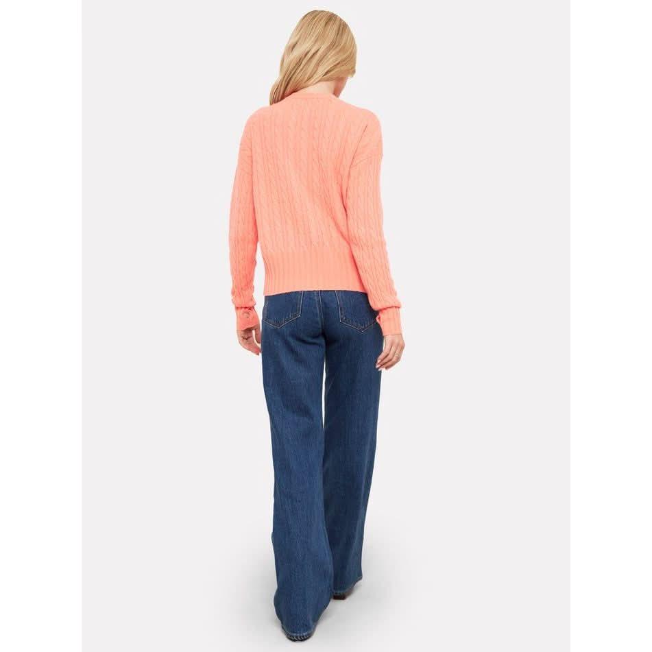 A person with long hair is wearing the Brodie Cashmere Cathy Cashmere Cable Knit sweater in a peach color and blue wide-leg jeans. They are facing away from the camera, walking on a white background.