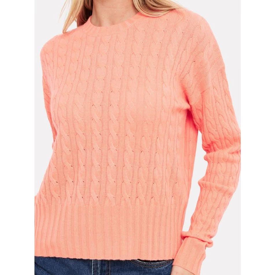 A person wearing the Brodie Cashmere Cathy Cashmere Cable Knit in neon coral pairs it with blue jeans against a plain background. This sweater from Brodie Cashmere boasts a textured cable-knit pattern, accompanied by ribbed cuffs and a ribbed crew neckline, adding a touch of elegance to this casual ensemble.