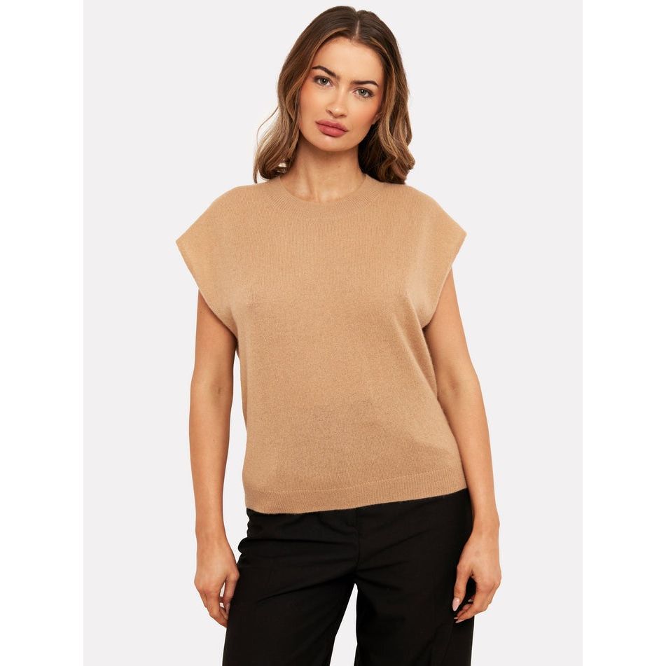 A person with long hair is wearing the Brodie Cashmere Tabby Cashmere Tank in Camel along with black pants, standing against a plain white background. They strike a relaxed pose with one hand slightly on their hip.