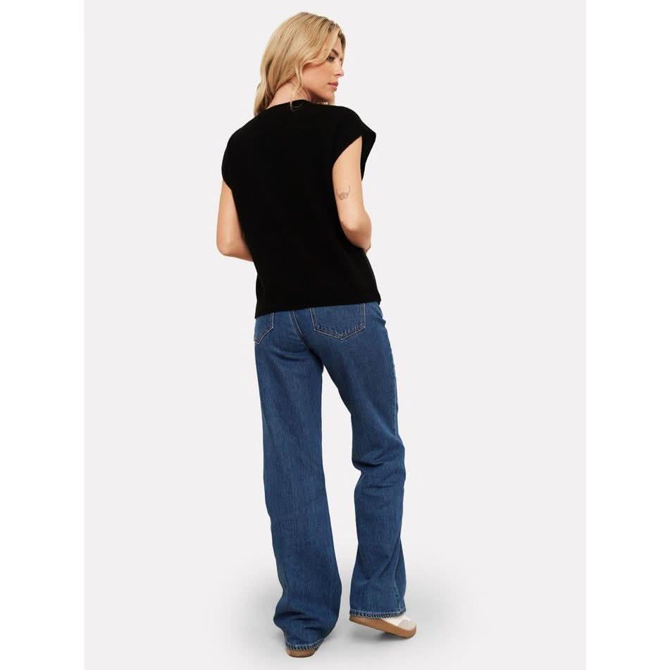 A person with long blonde hair stands with their back to the camera, wearing a machine washable Brodie Cashmere Tabby Cashmere Tank in black and blue jeans. They glance over their shoulder against a plain white background.