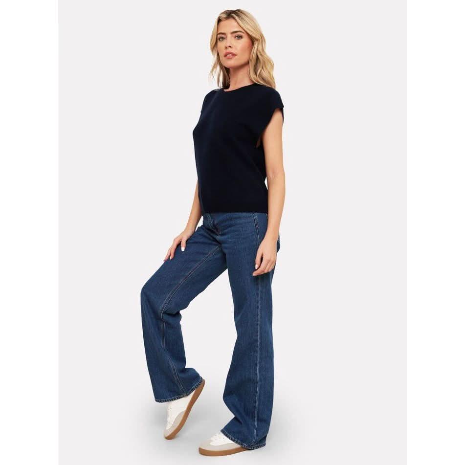 A person poses confidently in a Brodie Cashmere Tabby Cashmere Tank in black, wide-leg blue jeans, and white sneakers. The ensemble is elevated by the luxurious and cozy touch of cashmere against the plain white background.