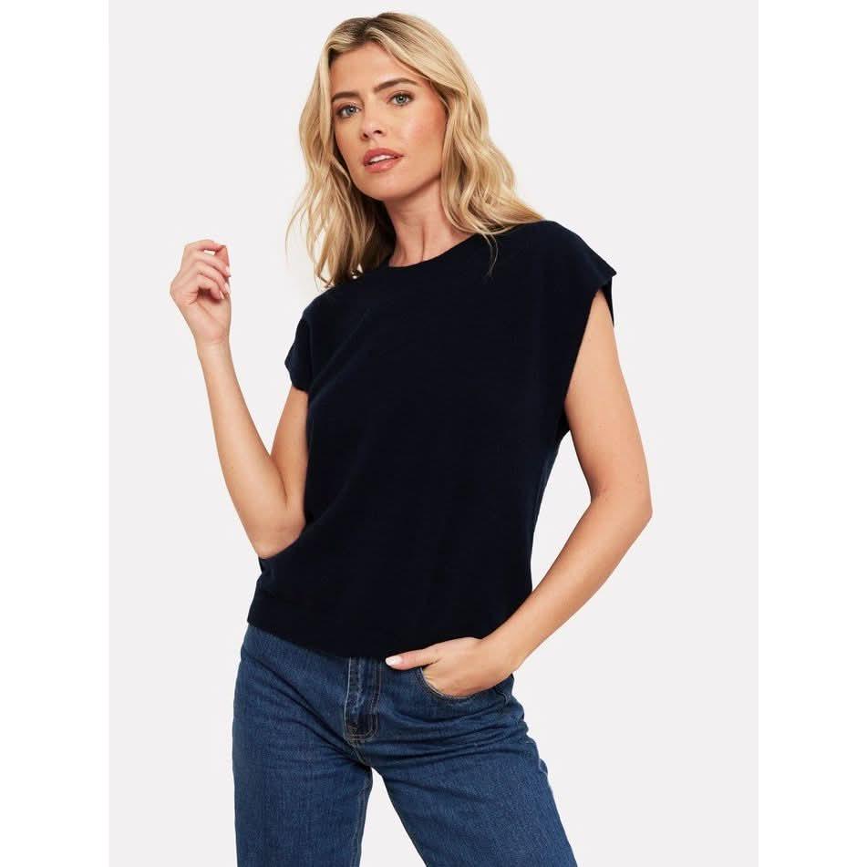 A woman with long, wavy blonde hair is wearing the Brodie Cashmere Tabby Cashmere Tank in black and blue jeans. She poses with one hand in her pocket and the other slightly raised, exuding effortless elegance against the plain white backdrop—an image as classic and versatile as the luxurious cashmere from Brodie Cashmere.