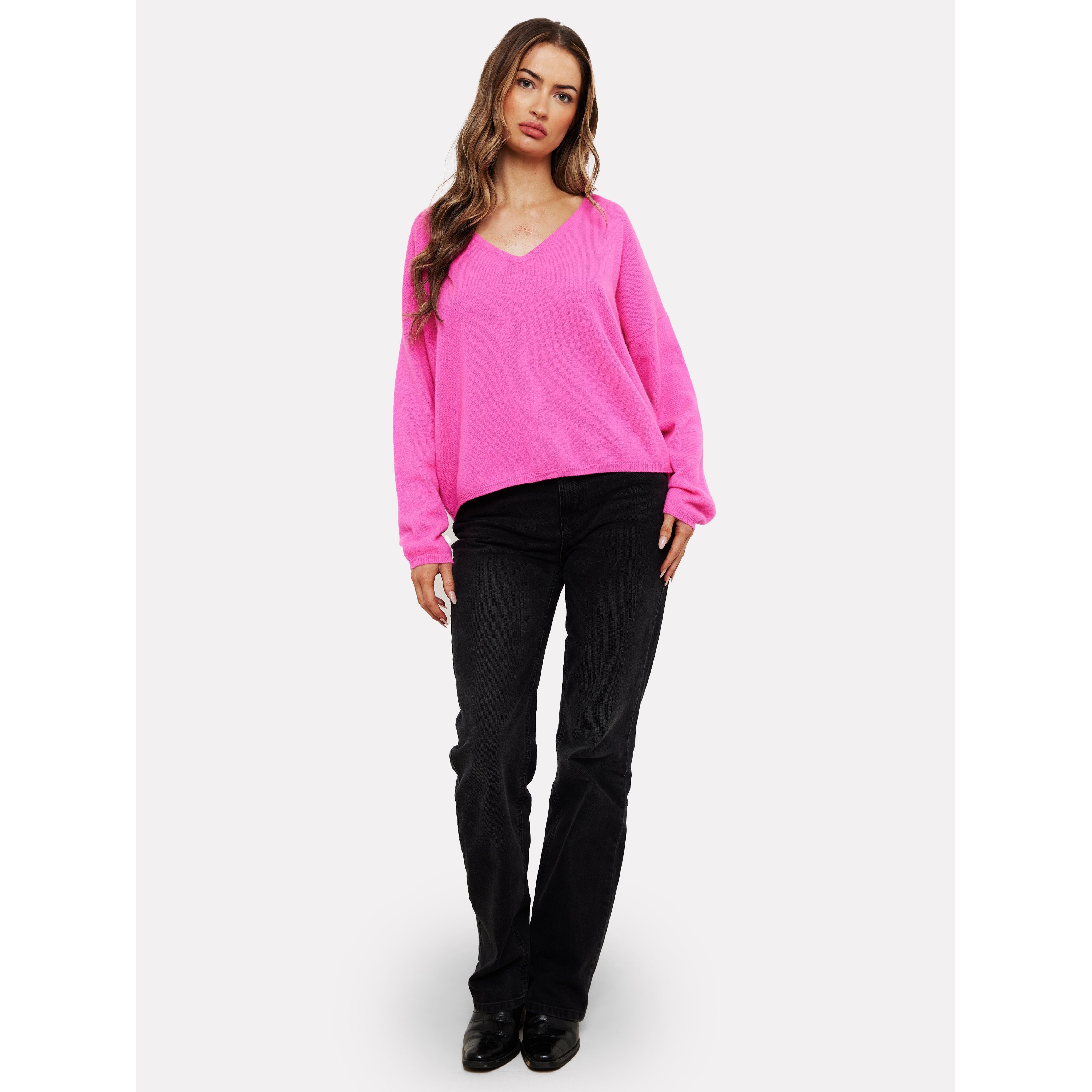 A woman with long wavy hair leans casually against a plain background wearing the vibrant Brodie Cashmere Rosa Reversible Diva Pink sweater, elegantly paired with black jeans and shoes. Her relaxed posture exudes effortless style while the luxurious Mongolian cashmere ensures comfort with its non-toxic dyes.