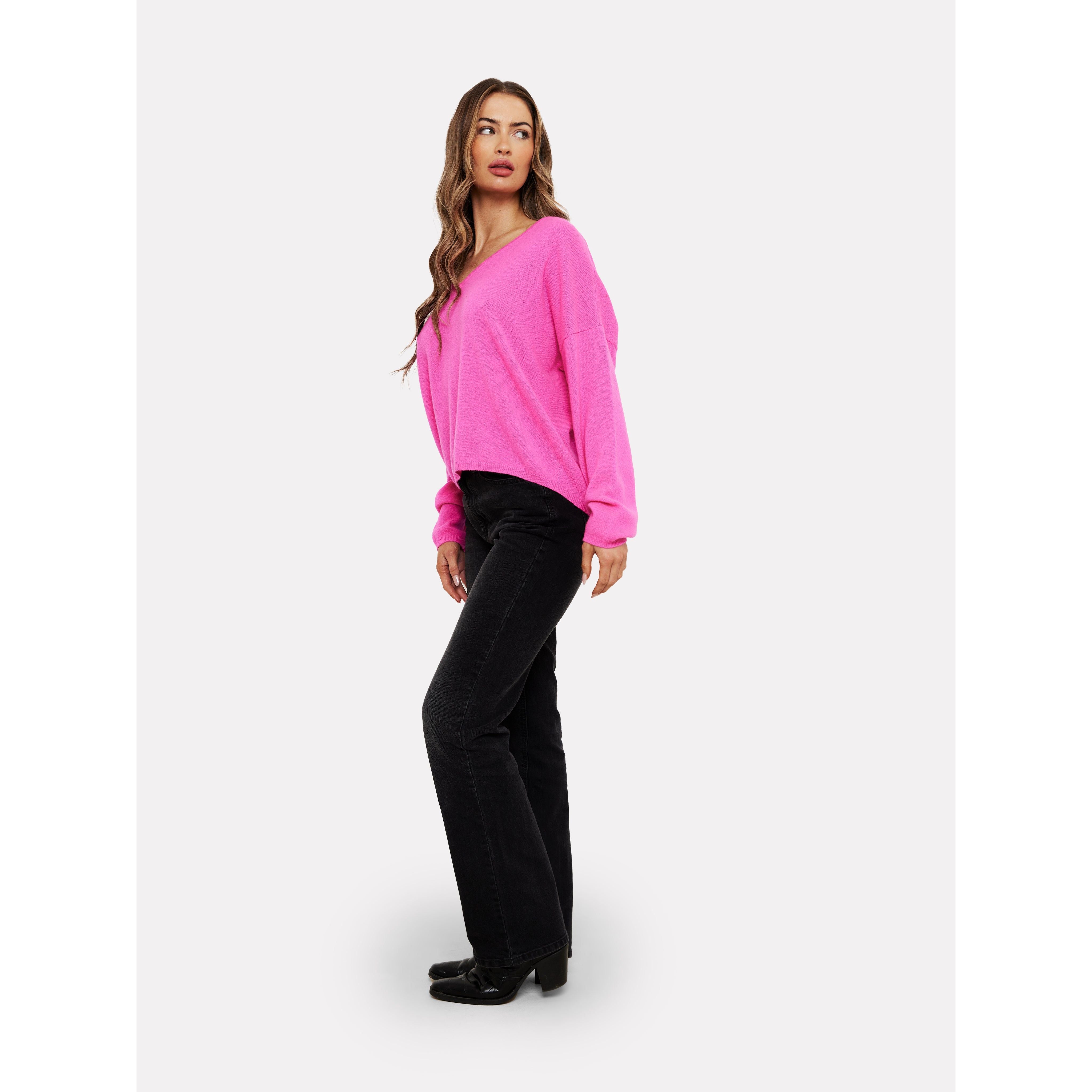 A person with long hair stands against a plain background, wearing the vibrant Brodie Cashmere Rosa Reversible Diva Pink sweater, complemented by black pants and shoes. They are slightly turned to the side and gazing into the distance.