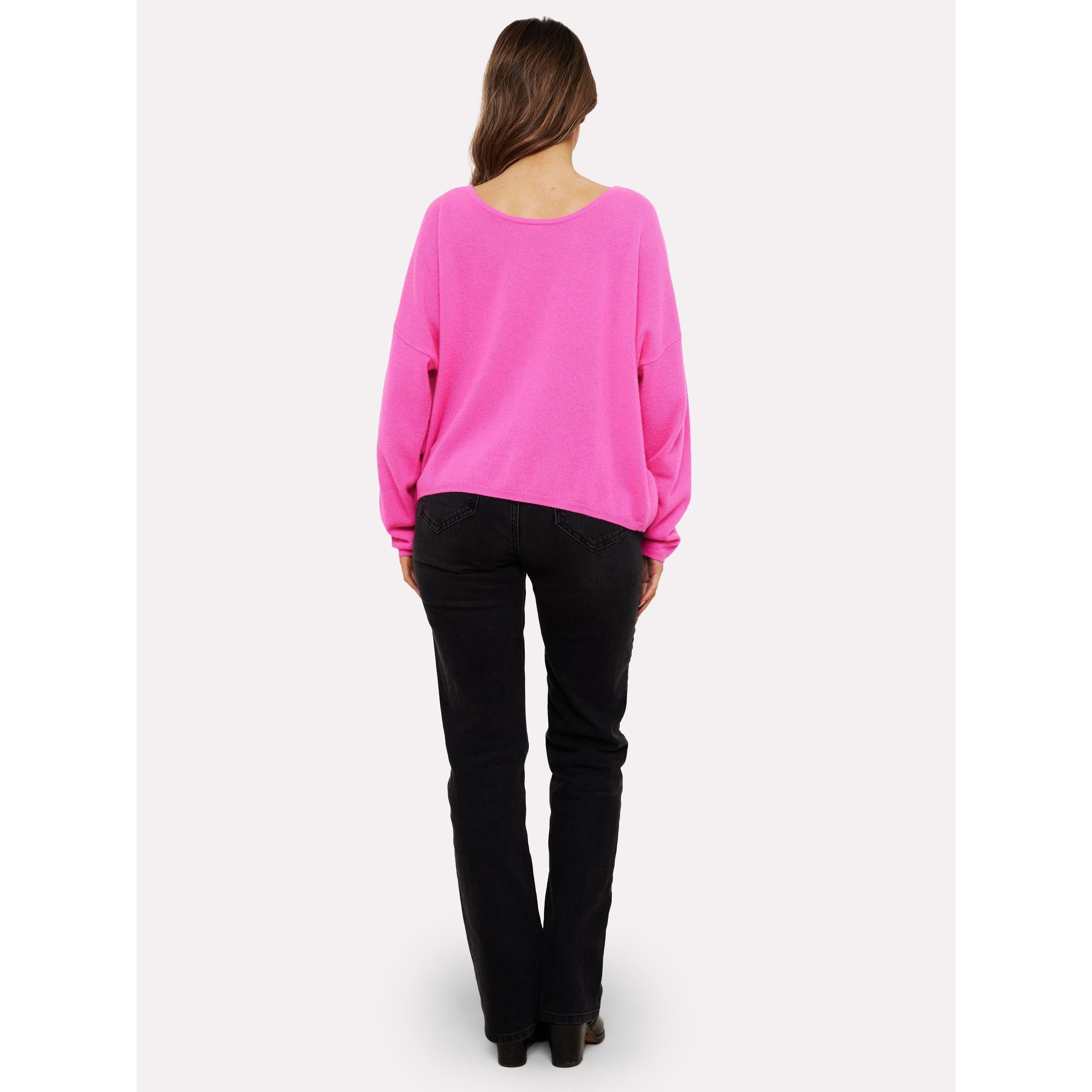 Facing away, a person with long hair showcases the Brodie Cashmere Rosa Reversible Diva Pink sweater and black jeans, set against a pristine white background.