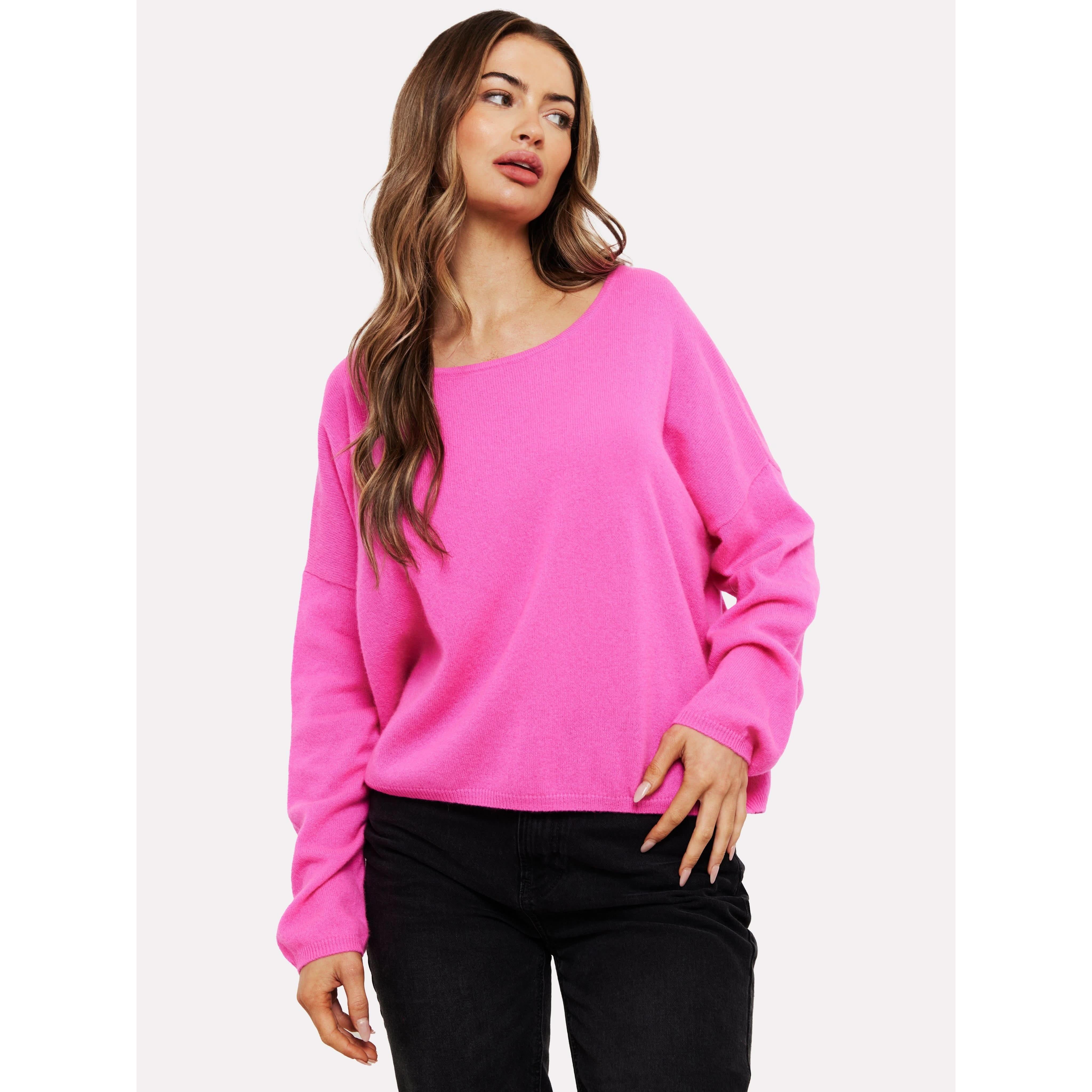 A person with long, wavy hair wears the Brodie Cashmere Rosa Reversible Diva Pink sweater from Brodie Cashmere, made with non-toxic dyes, paired with black pants. They look to the side with a neutral expression, their right hand resting near the hem of the sweater against a plain white background.