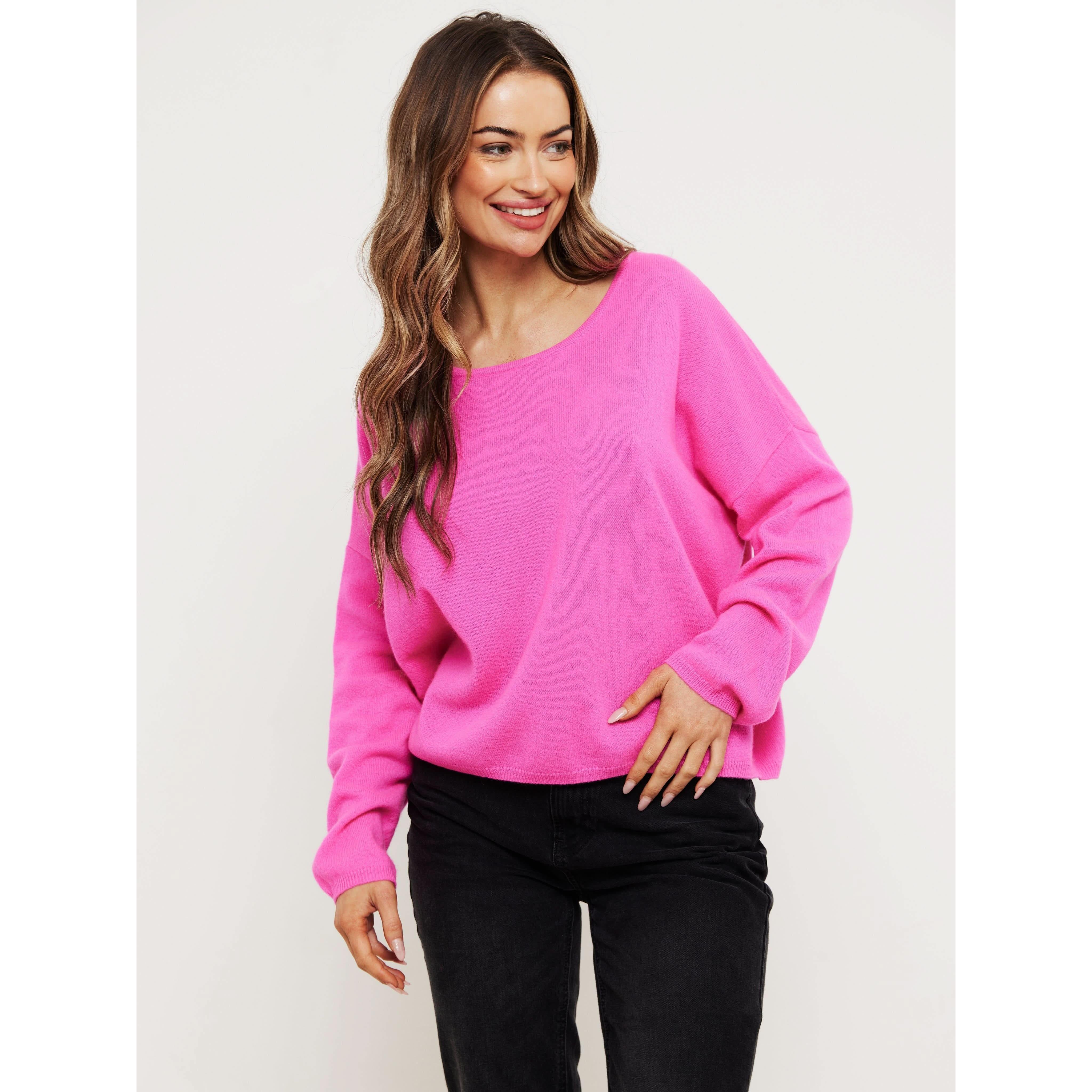 A person with long hair is smiling and wearing the Brodie Cashmere Rosa Reversible Diva Pink sweater, made from Mongolian cashmere and featuring non-toxic dyes, complemented by black pants. The vibrant pink sweater stands out against the plain white background.