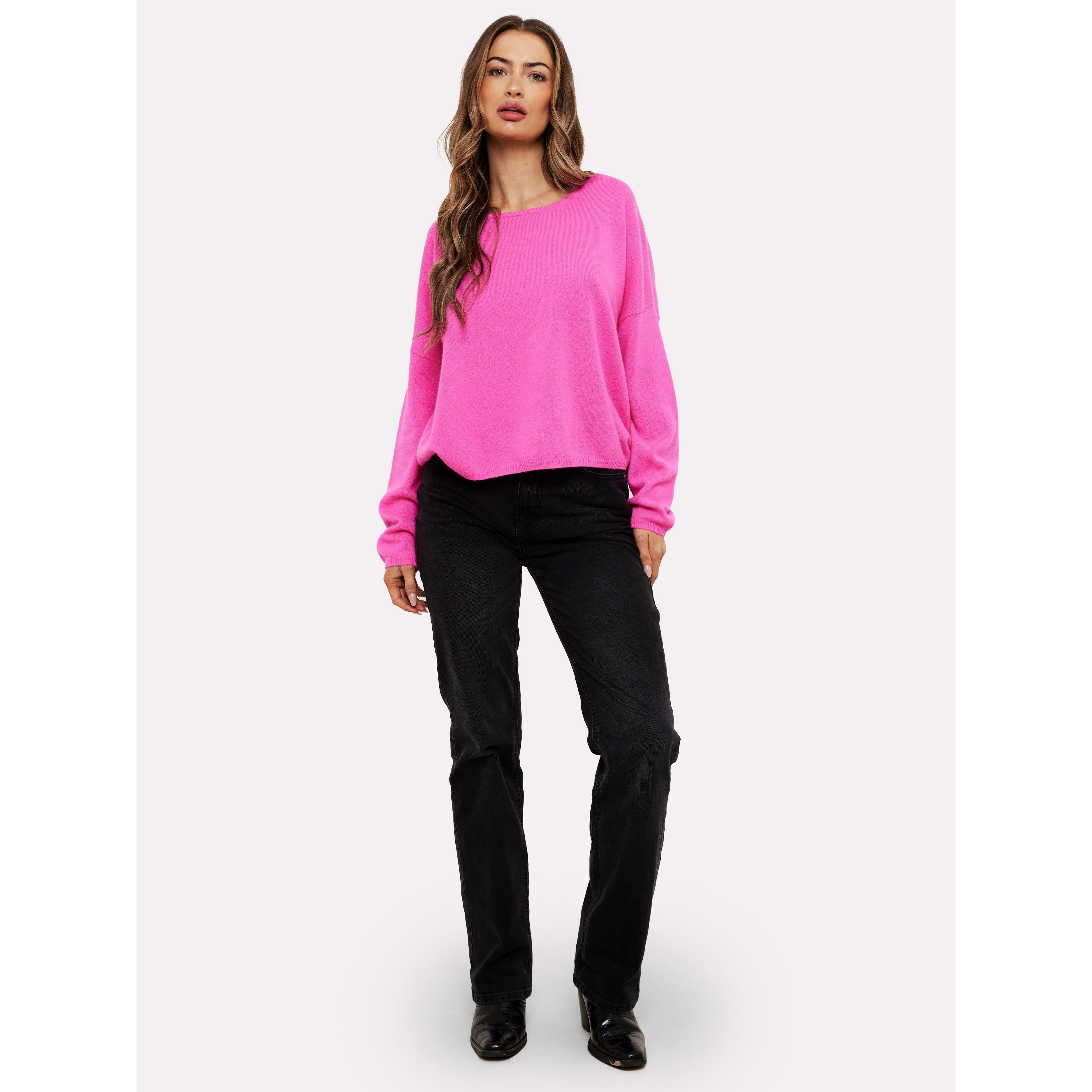 A person wearing the vibrant Brodie Cashmere Rosa Reversible Diva Pink sweater, crafted from Mongolian cashmere and machine washable, pairs it with black jeans. They stand against a plain white background, have long hair, and wear black shoes dyed with non-toxic dyes.