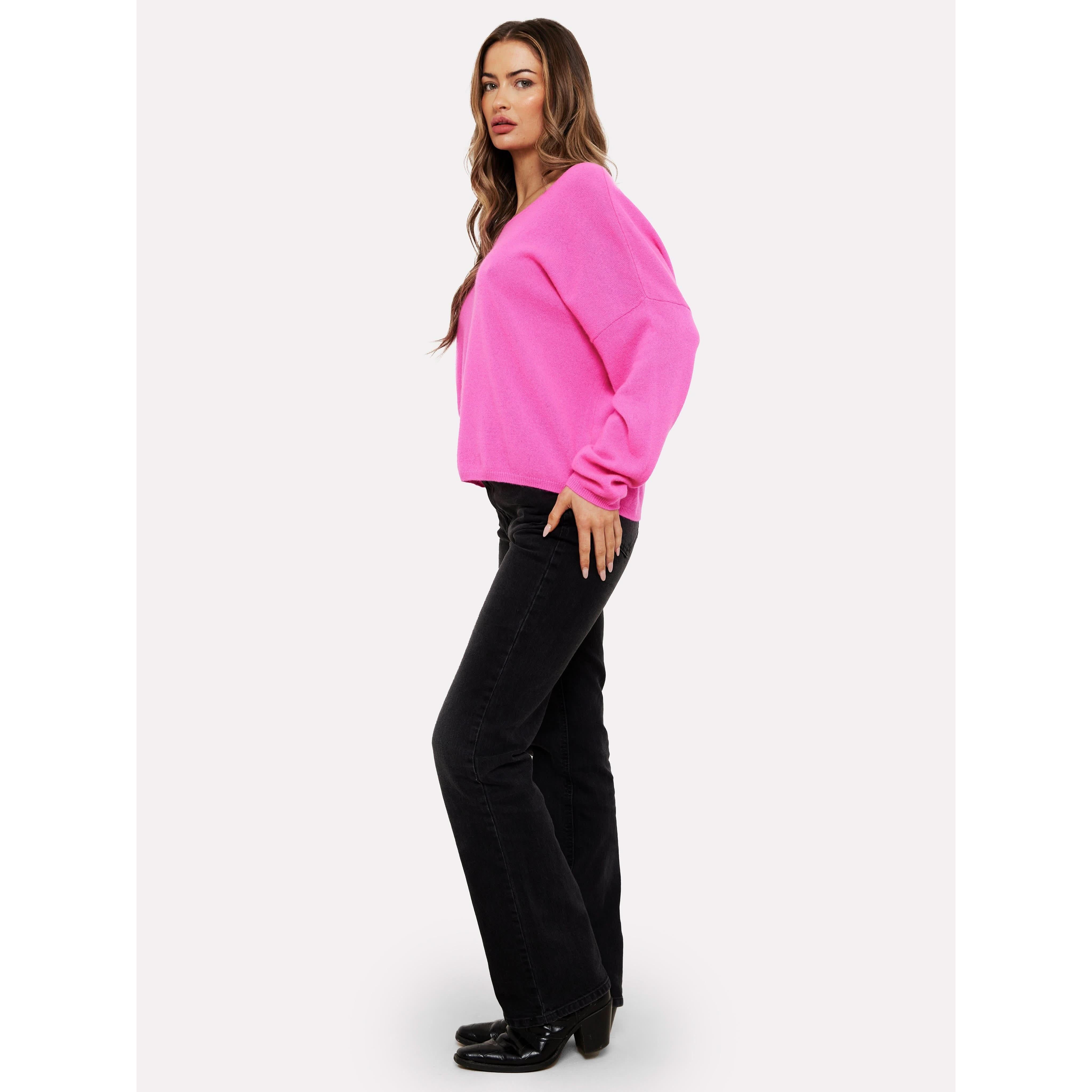 A person with long hair stands sideways, wearing a Brodie Cashmere Rosa Reversible Diva Pink sweater, dark flared jeans, and black shoes. They have a confident expression and are posing against a plain white background, effortlessly stylish in attire that's as luxurious as it is machine washable.