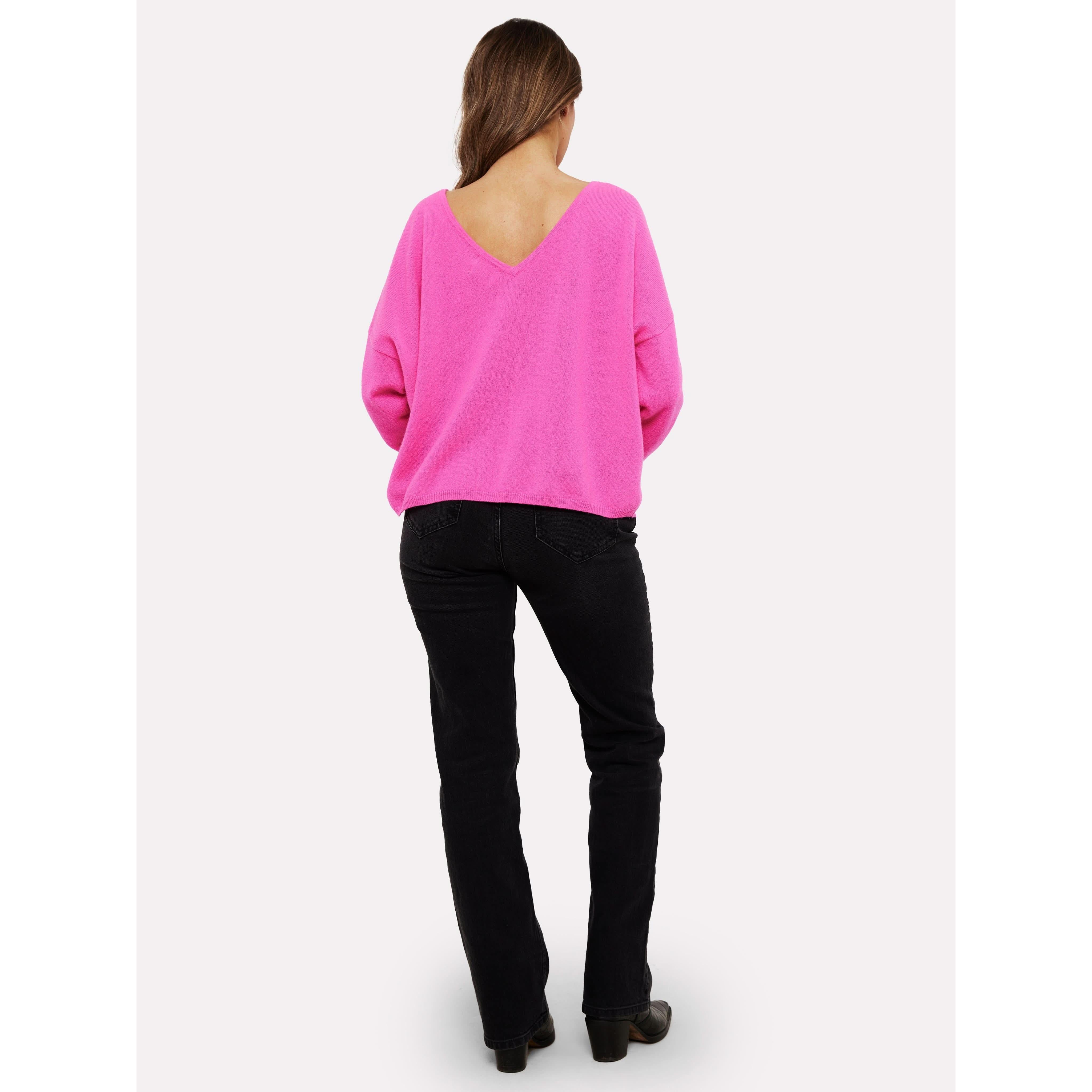 A person with long hair stands with their back to the camera, highlighting the Brodie Cashmere Rosa Reversible Diva Pink sweater, known for its vibrant pink hue and V-shaped back, crafted using non-toxic dyes. They have effortlessly paired it with sleek black pants.