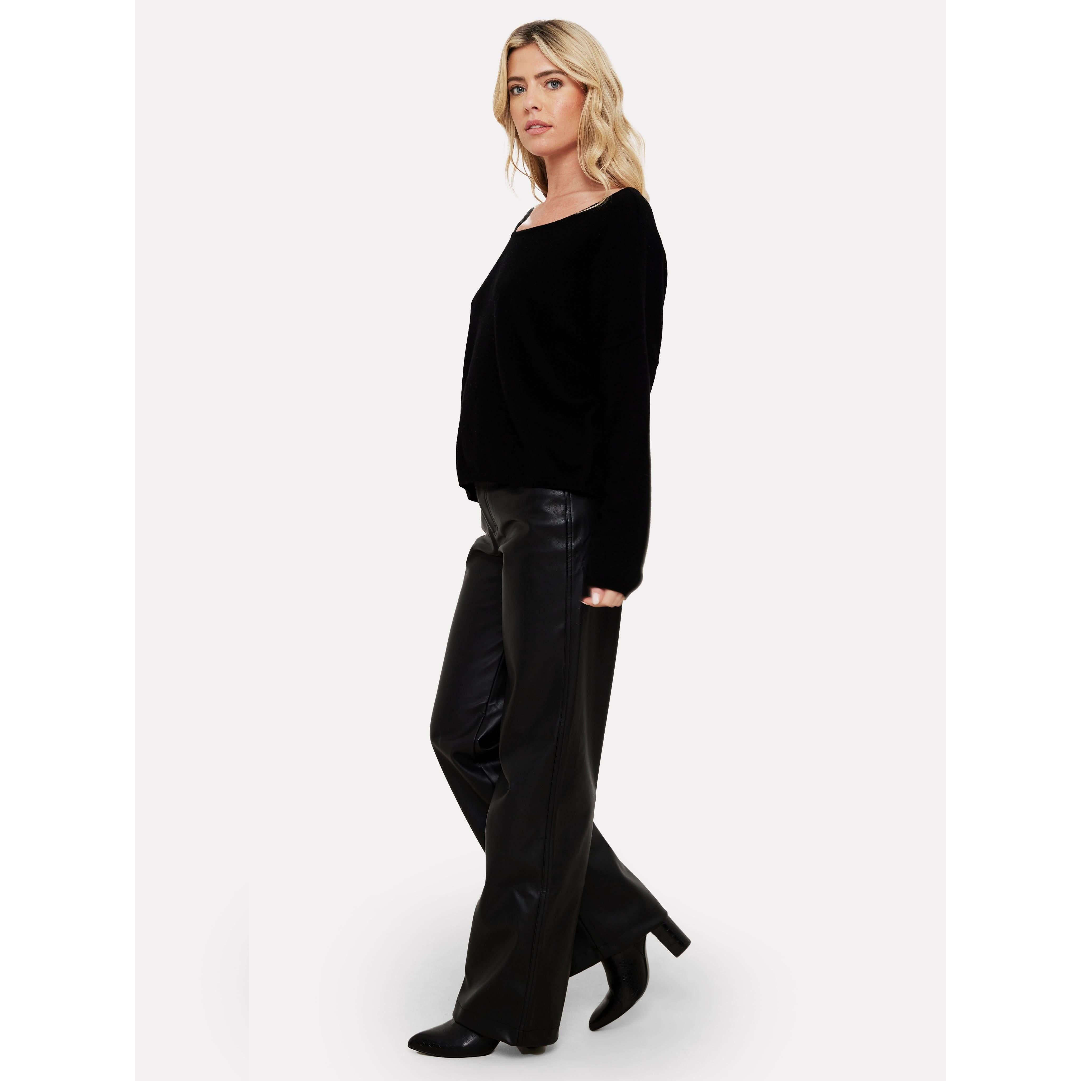 A person wearing the Brodie Cashmere Rosa Reversible Black sweater and black leather pants stands against a plain white background. They have long blonde hair and are looking off to the side, complemented by stylish black heeled shoes from Brodie Cashmere.