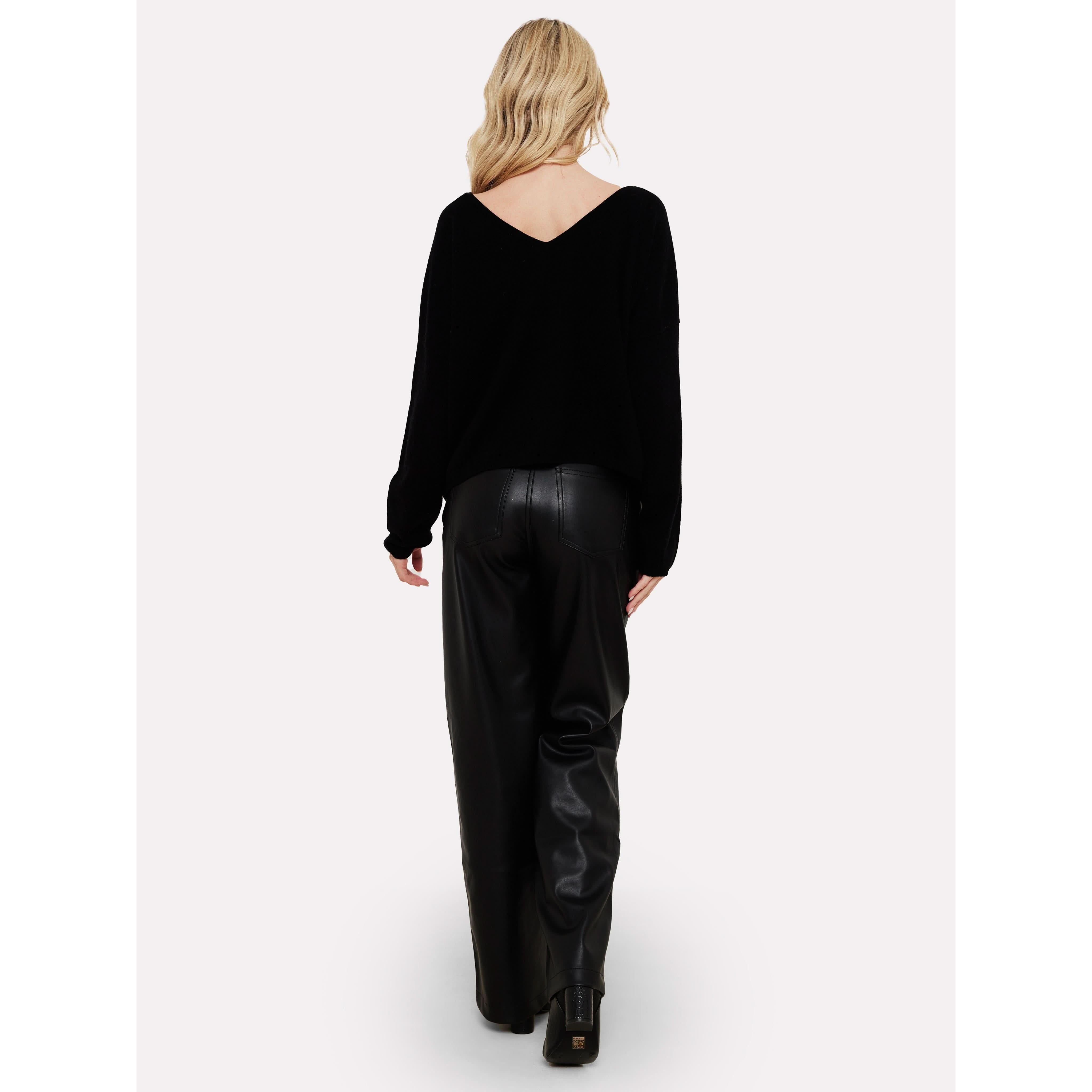 Rear view of a person with long, blonde hair wearing the Brodie Cashmere Rosa Reversible Black sweater from Brodie Cashmere, wide-leg black leather pants, and black platform shoes, walking against a plain white background.