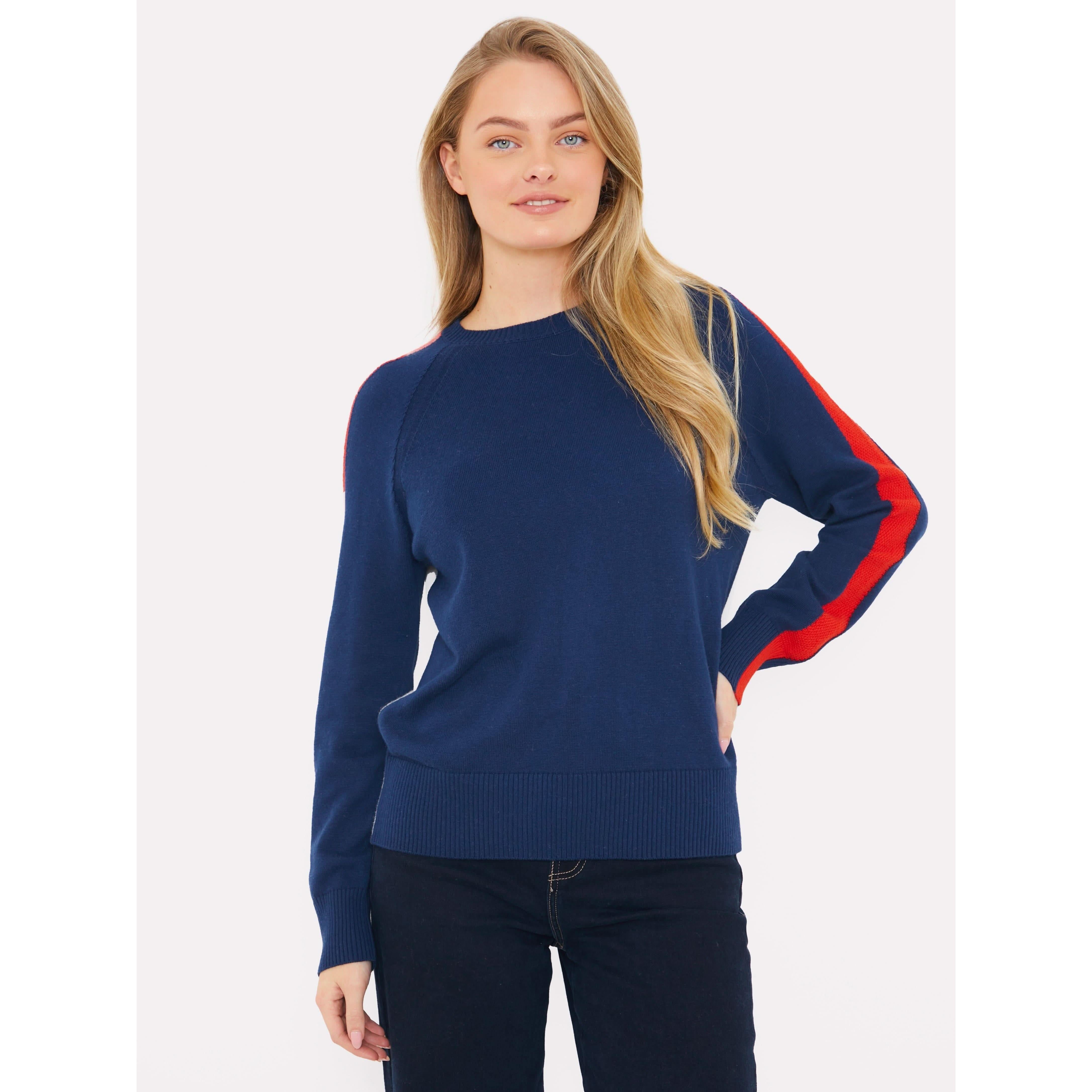 Someone with long hair is wearing the Brodie Cashmere Stripe Up Crew by Brodie Cashmere. This navy blue sweater features red stripes on the sleeves and thumb hole details. They pair it with dark pants while standing against a plain white background, subtly highlighting the unique design of the sweater.