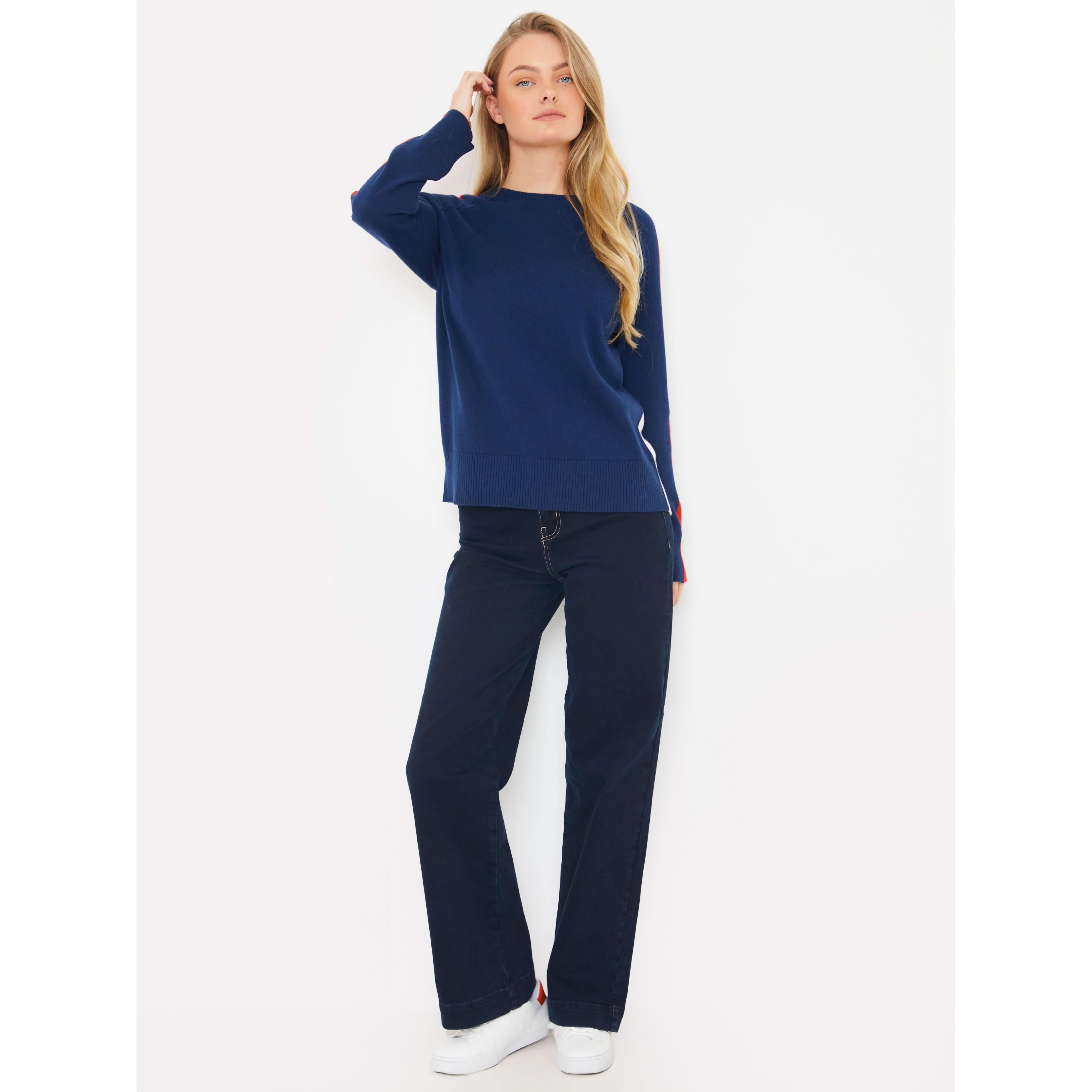 A woman stands against a white background wearing the Brodie Cashmere Stripe Up Crew sweater in navy blue, paired with dark jeans. With her long blonde hair and white sneakers, she presents a casual yet stylish look that effortlessly blends comfort and fashion.