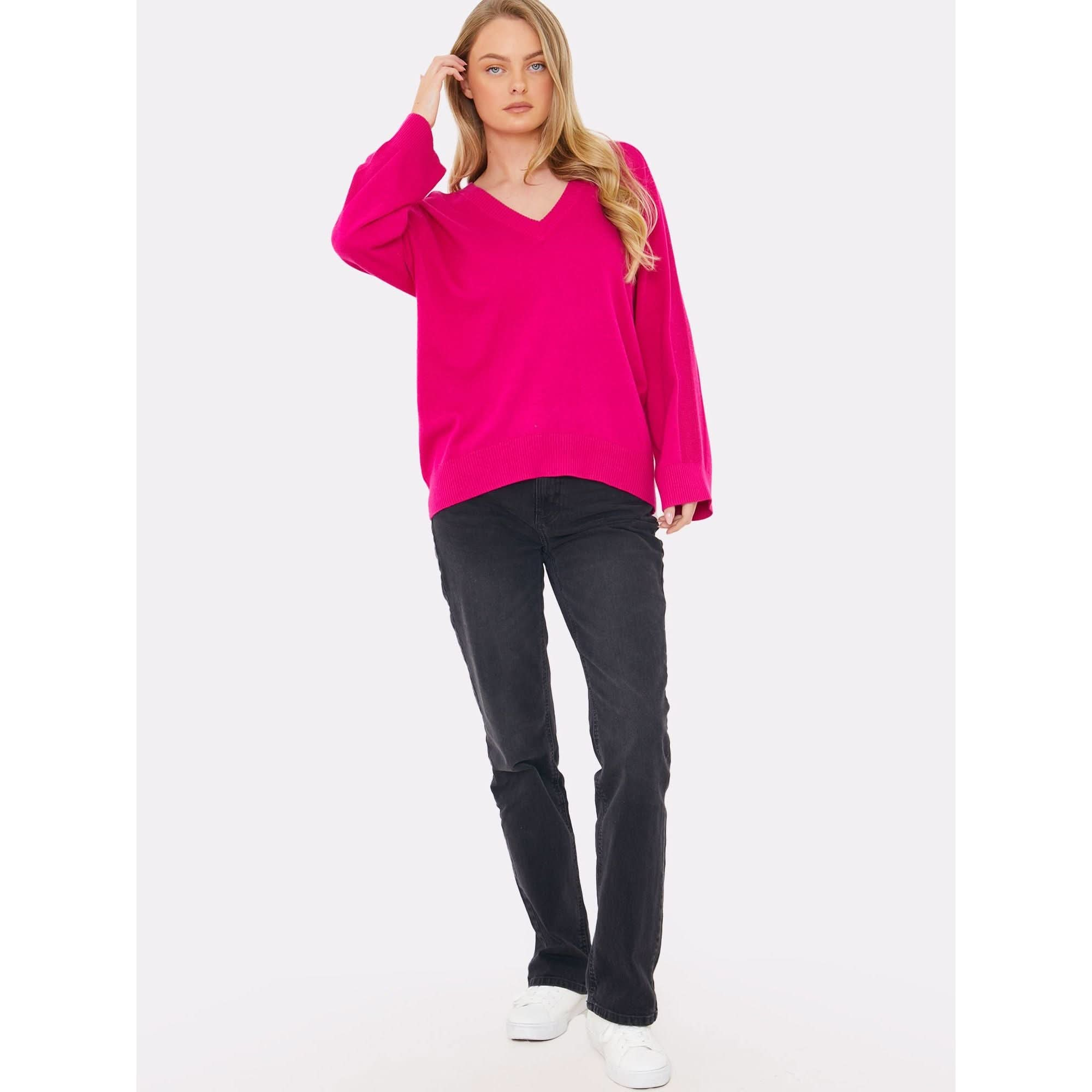 A person with long blond hair is wearing the Brodie Cashmere Saddle Shoulder Rib Knit in fuschia from Brodie Cashmere, paired with black jeans and white sneakers. They are standing against a plain white background with one hand touching their head.