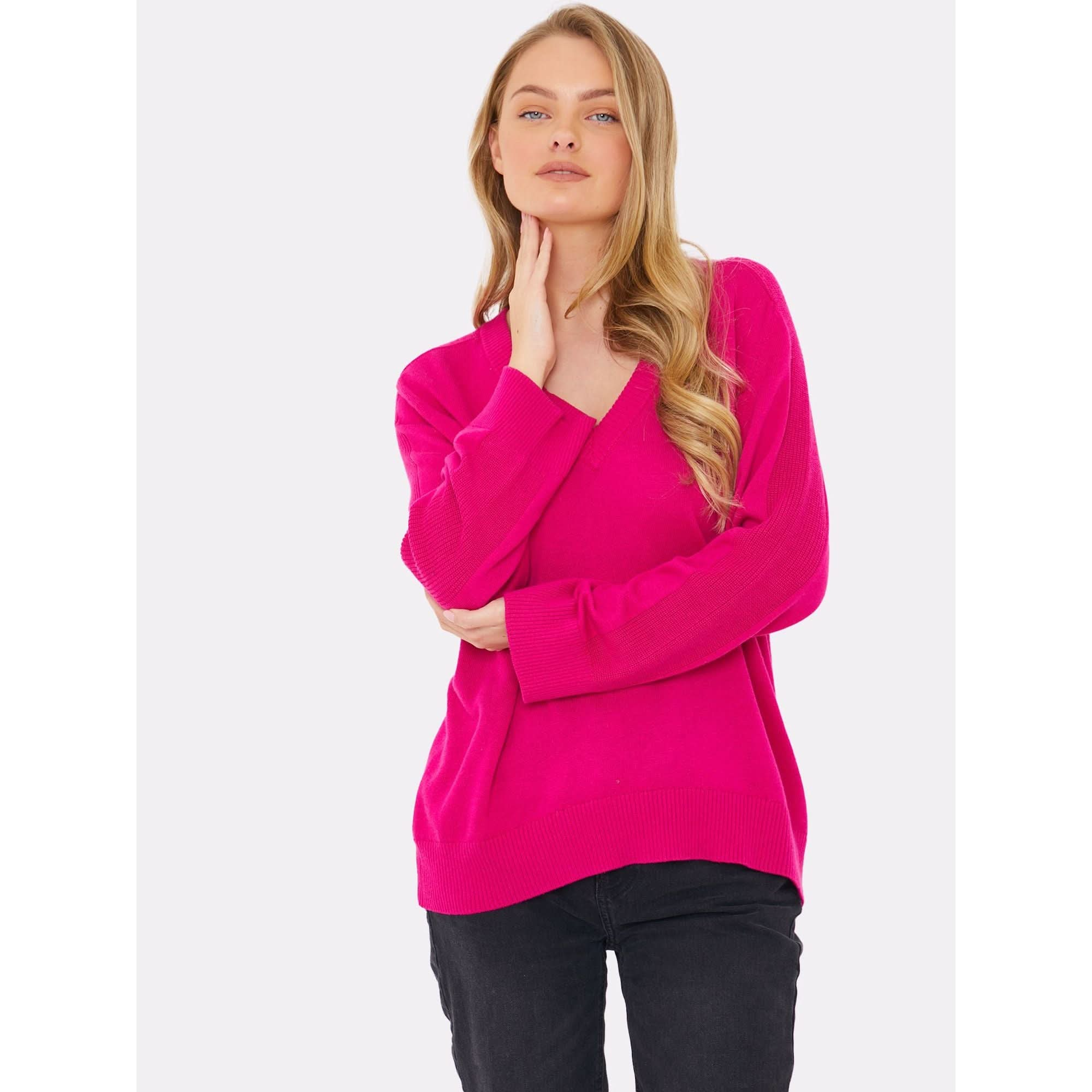 A person with long blonde hair, clad in a vibrant fuchsia sweater from the Brodie Cashmere Saddle Shoulder Rib Knit collection and black pants, poses with their hand on their chin against a plain white background. The sweater features a crew neckline and is crafted using a non-toxic dyeing method.