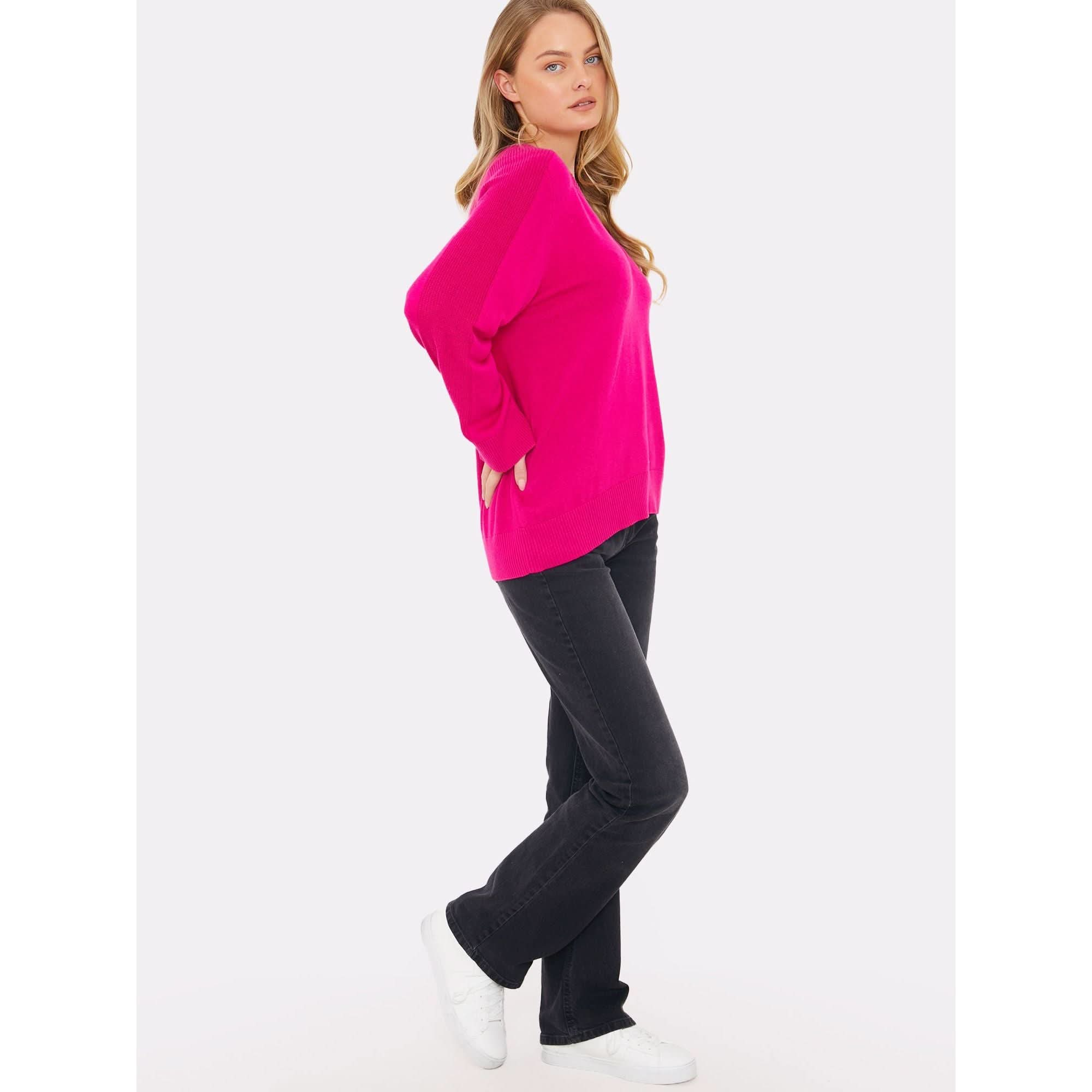 A person with long hair stands sideways, wearing a vibrant fuschia sweater from the Brodie Cashmere collection featuring saddle shoulder rib knit design and a crew neckline, paired with black pants and white sneakers. Their hands are in their pockets, conveying a relaxed expression against a plain white background.