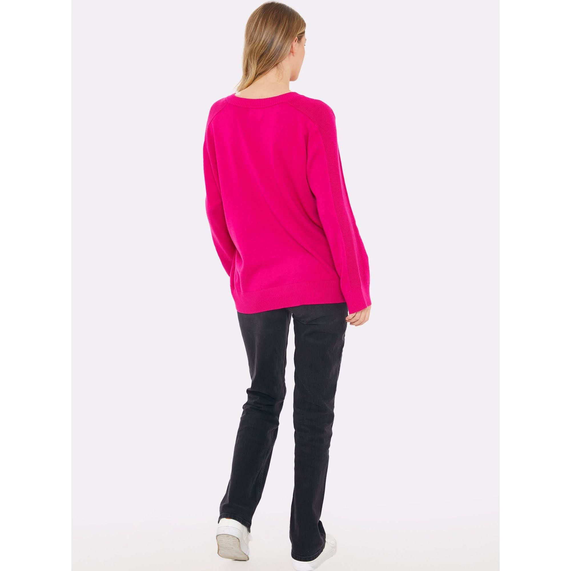 A person, dressed in the Brodie Cashmere Saddle Shoulder Rib Knit Fuschia sweater from the Brodie Cashmere collection, is wearing black pants and white shoes. They are standing and facing away from the camera against a plain white background.