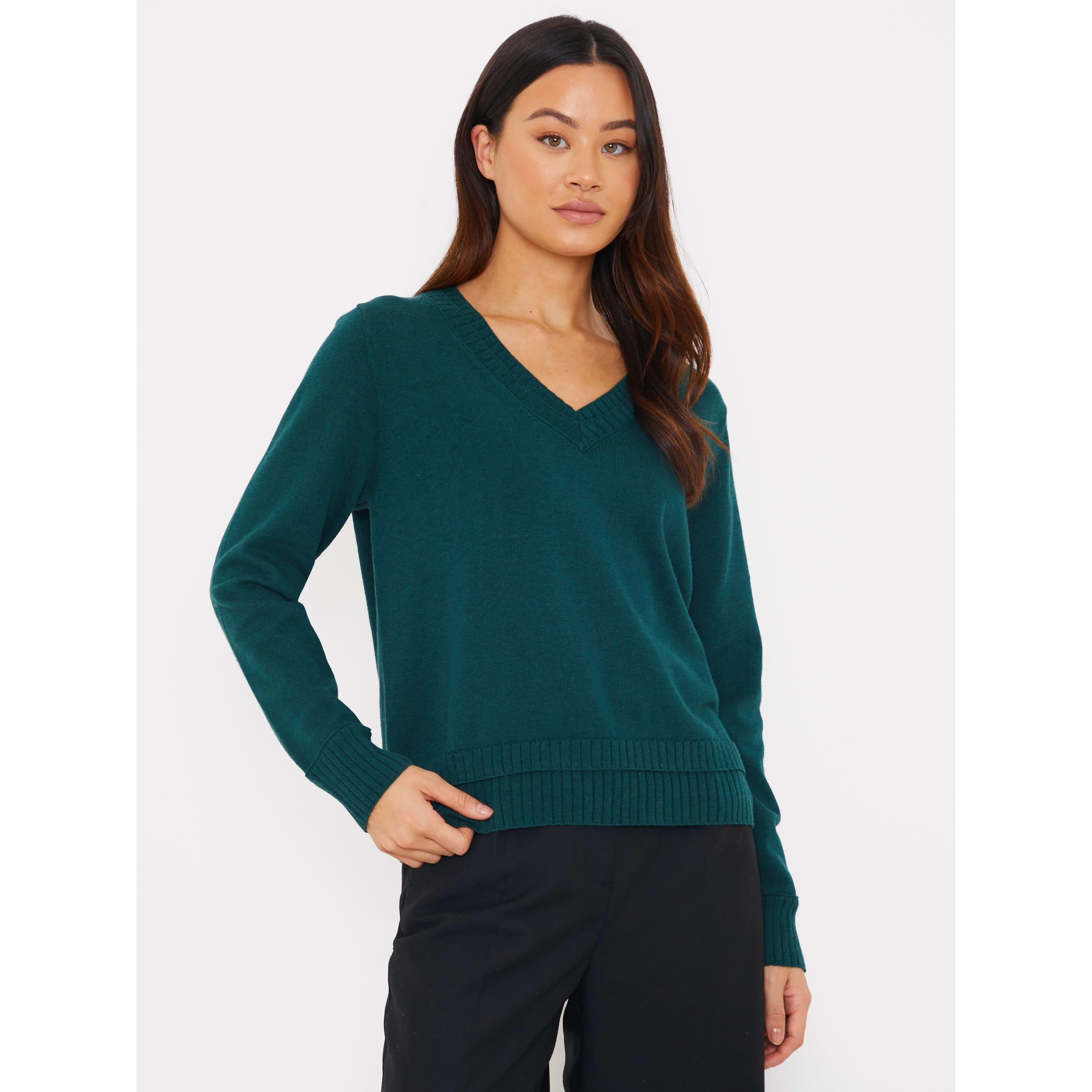 A person with long dark hair wears the Brodie Cashmere Double Welt V Neck sweater in a deep green shade, paired with black pants. They stand against a plain white background.