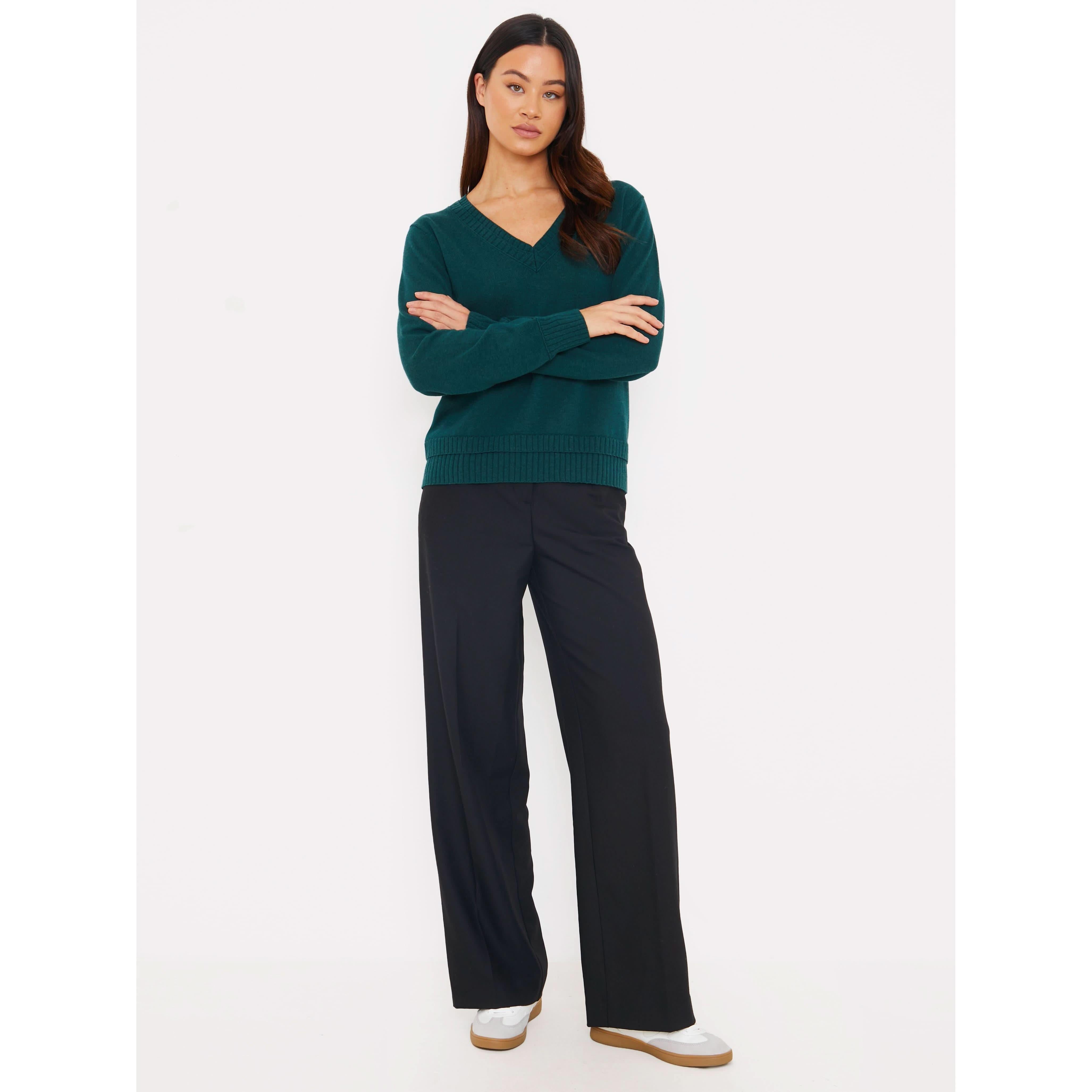 Against a plain white background, a person wears the Brodie Cashmere Double Welt V Neck sweater in dark green, paired with black wide-leg pants and white sneakers. They have long dark hair and pose with arms crossed, looking directly at the camera.