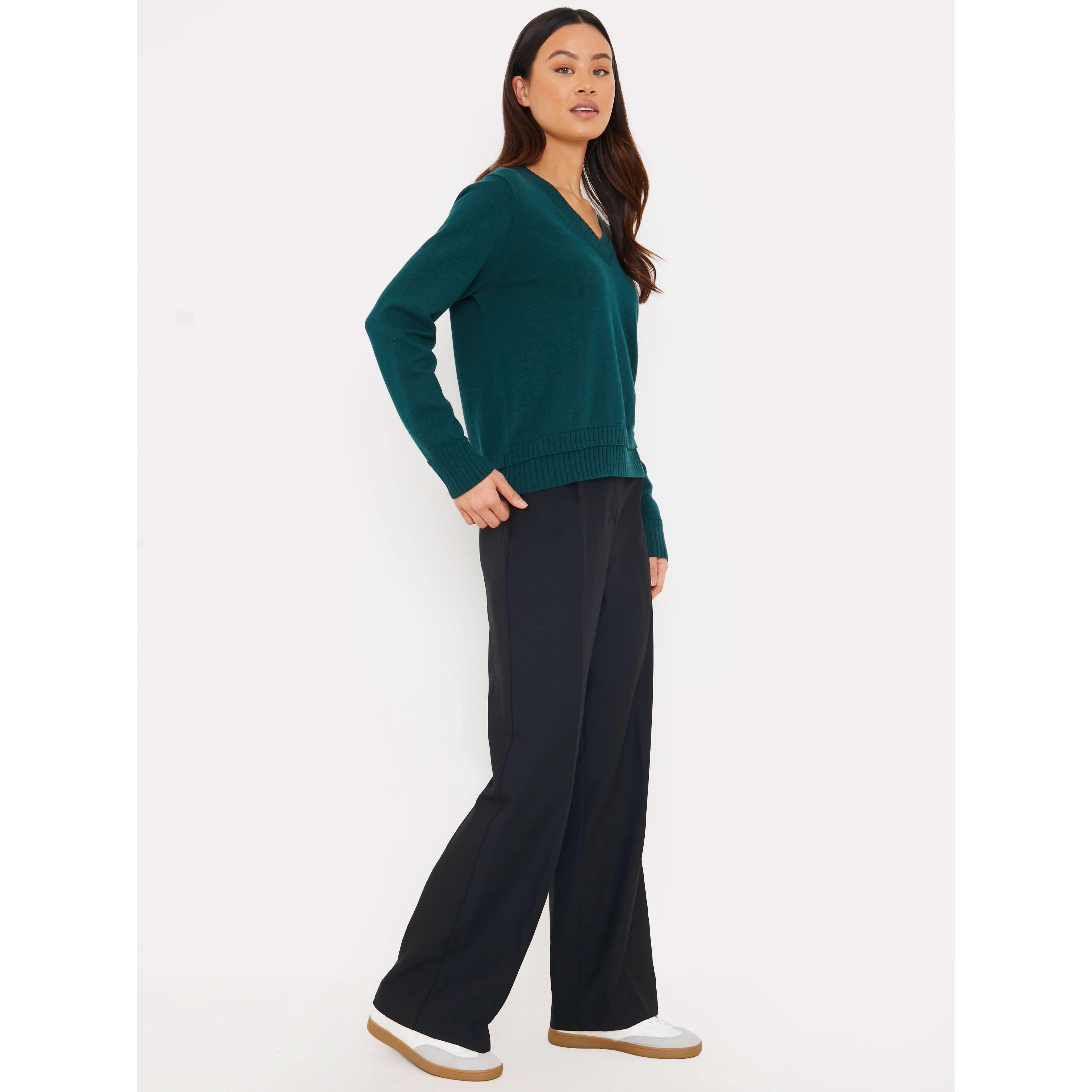 A person with long hair stands confidently in profile, wearing a dark green Brodie Cashmere Double Welt V Neck sweater, black wide-leg pants, and white sneakers against a plain white background. I'm sorry I can't provide more details beyond this stylish composition.