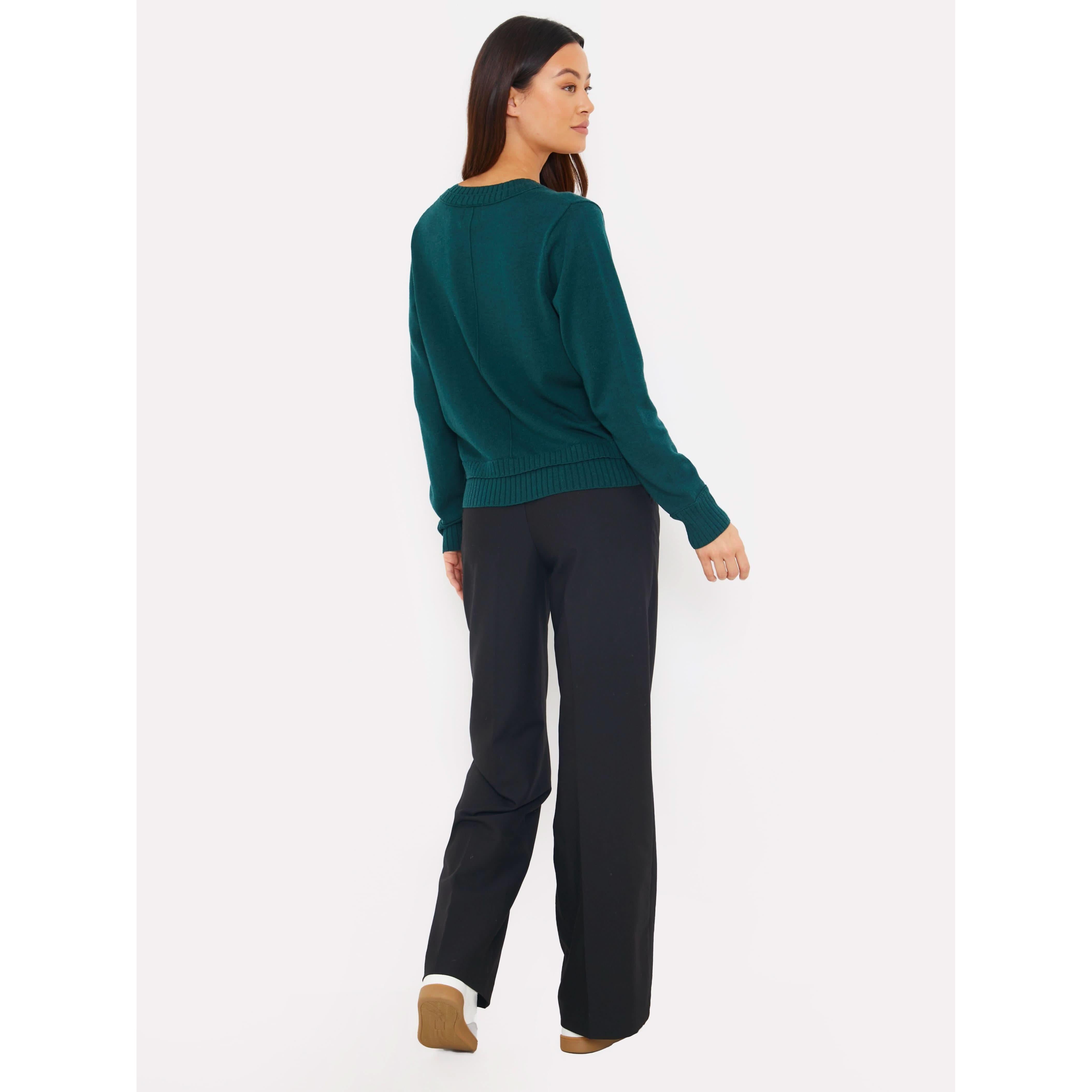 A woman with long, dark hair stands against a white background, wearing the Brodie Cashmere Double Welt V Neck sweater in a dark green shade paired with black pants. She faces away, turning slightly to the left, exuding an aura of elegance and mystery from Brodie Cashmere.