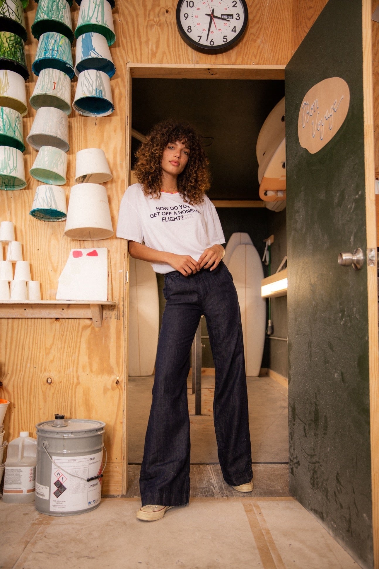 A woman with curly hair stands casually in a workshop, leaning against a door frame. She wears a white t-shirt and high-waisted ASKK NY Denim Trouser Indigo Linen pants. The workshop has various ceramic pieces on shelves, a pot of paint, and a clock on the wall.