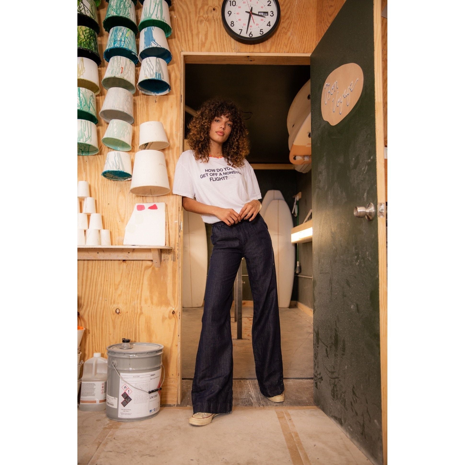 A woman with curly hair stands casually in a workshop, leaning against a door frame. She wears a white t-shirt and high-waisted ASKK NY Denim Trouser Indigo Linen pants. The workshop has various ceramic pieces on shelves, a pot of paint, and a clock on the wall.