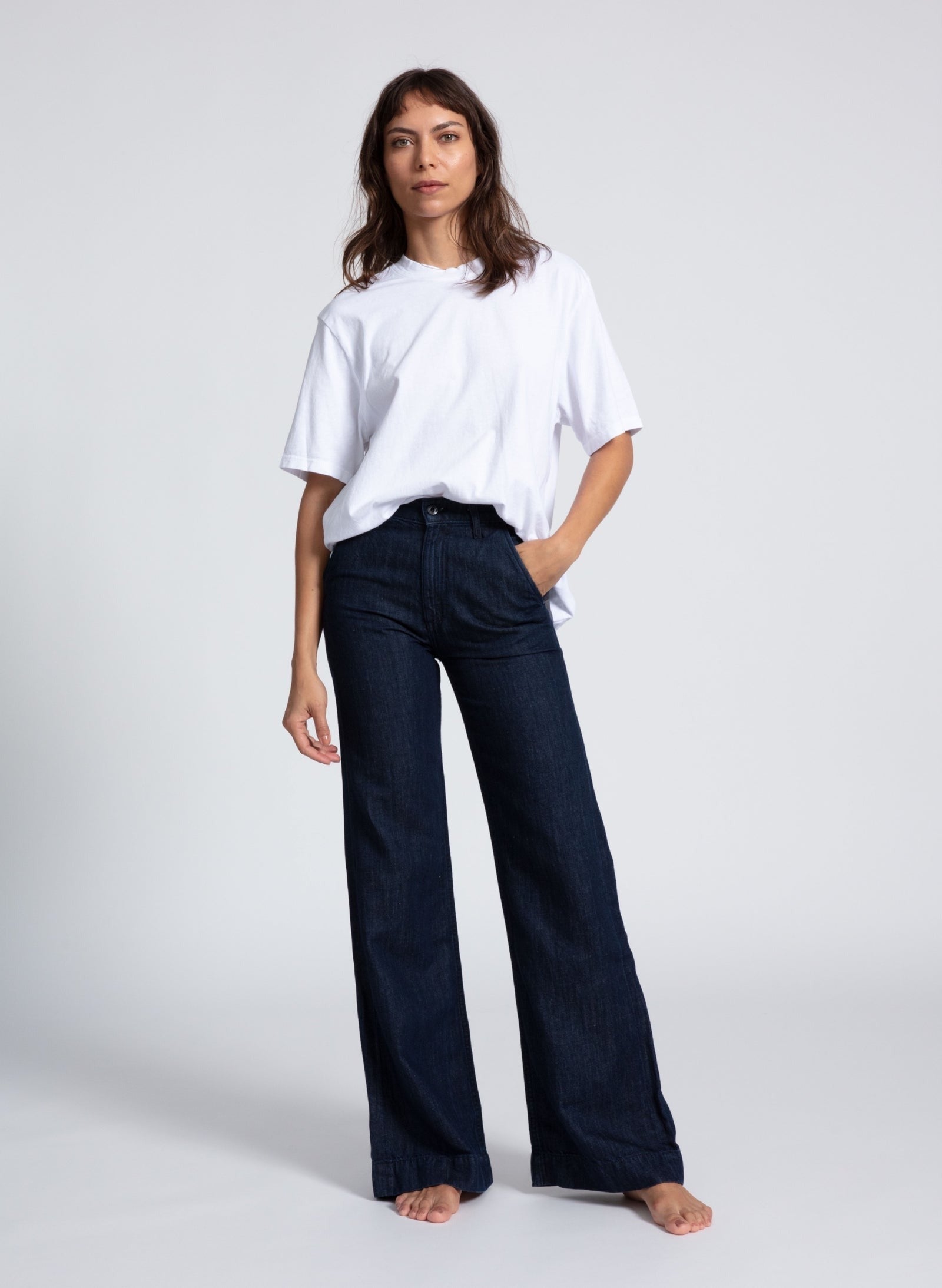 A person stands barefoot against a plain background, wearing a white, short-sleeved, loose-fitting t-shirt and the ASKK NY Denim Trouser Indigo Linen by ASKK NY. One hand is in their pocket while the other hangs by their side. Their expression is neutral.