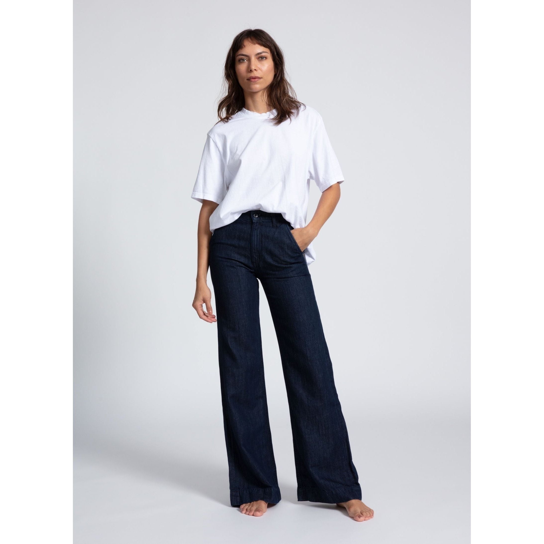 A person stands barefoot against a plain background, wearing a white, short-sleeved, loose-fitting t-shirt and the ASKK NY Denim Trouser Indigo Linen by ASKK NY. One hand is in their pocket while the other hangs by their side. Their expression is neutral.