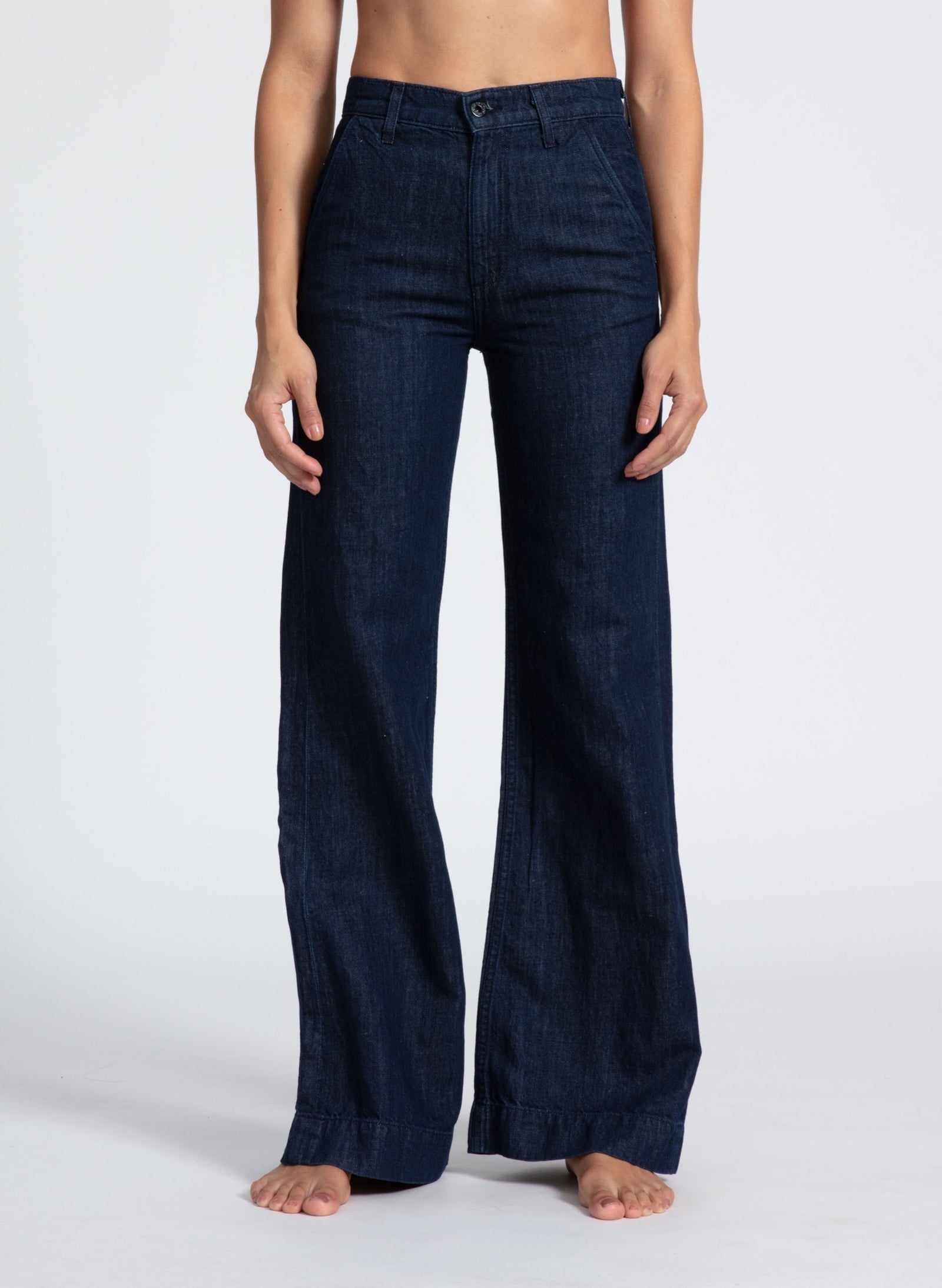 A person wearing the ASKK NY Denim Trouser in Indigo Linen stands barefoot against a plain white background. The high-waisted, wide-leg jeans, made by ASKK NY, feature a button and zipper closure. The person's hands rest by their sides as their upper body and feet are visible.