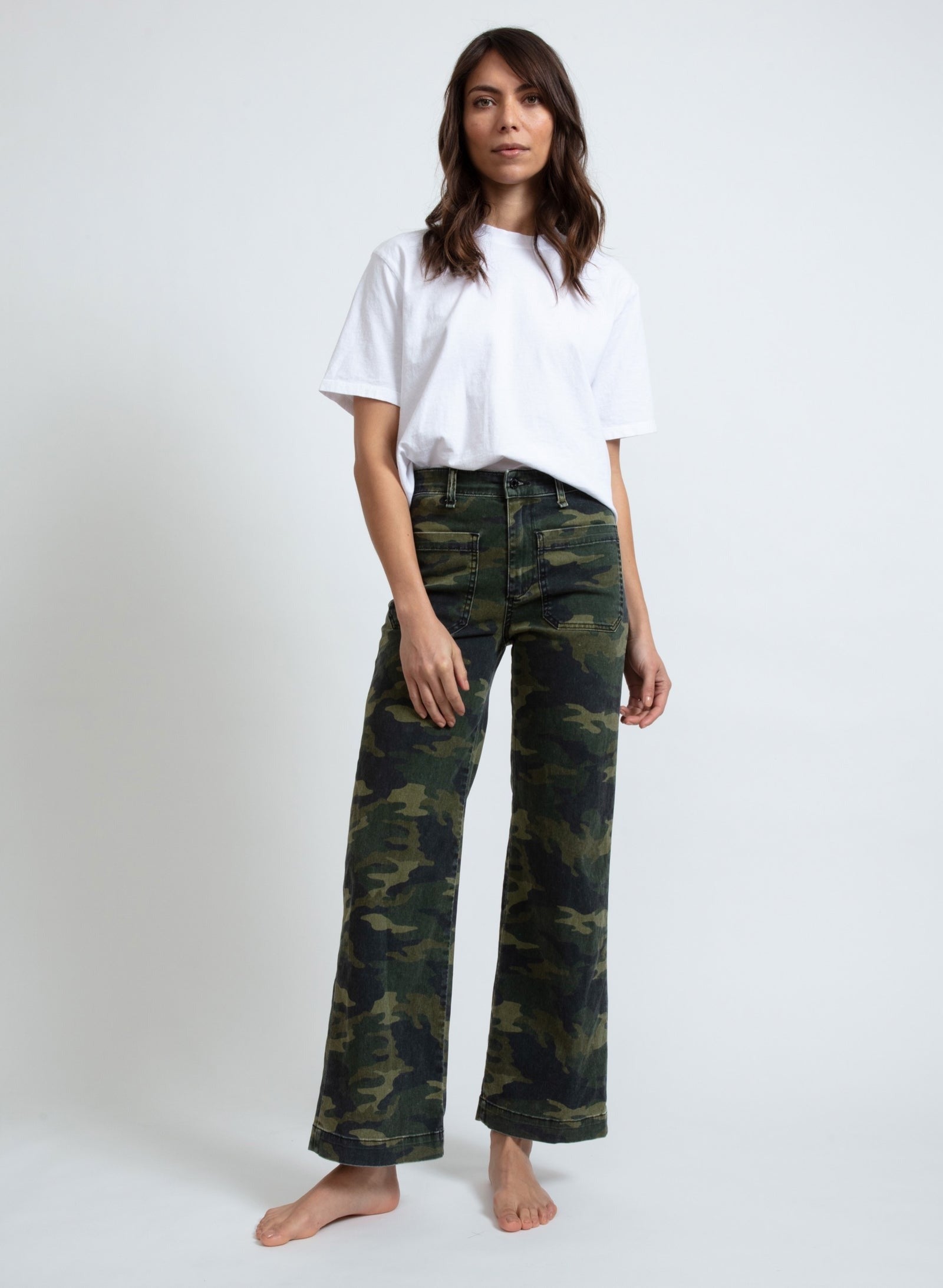 A person stands barefoot against a plain background, wearing a loose-fitting white T-shirt and high-waisted, wide-leg ASKK NY Printed Sailor Pants by ASKK NY. Their hands are relaxed at their sides, and they have long, wavy hair.