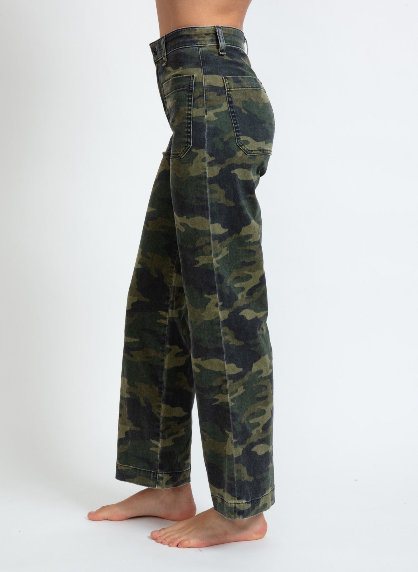 A person is standing sideways, wearing ASKK NY Printed Sailor Pant with a green and brown camouflage pattern. Made in Los Angeles by ASKK NY, these high-waisted pants feature a relaxed, straight-leg fit and have visible pockets on the backside. The person is barefoot, standing on a plain white background.