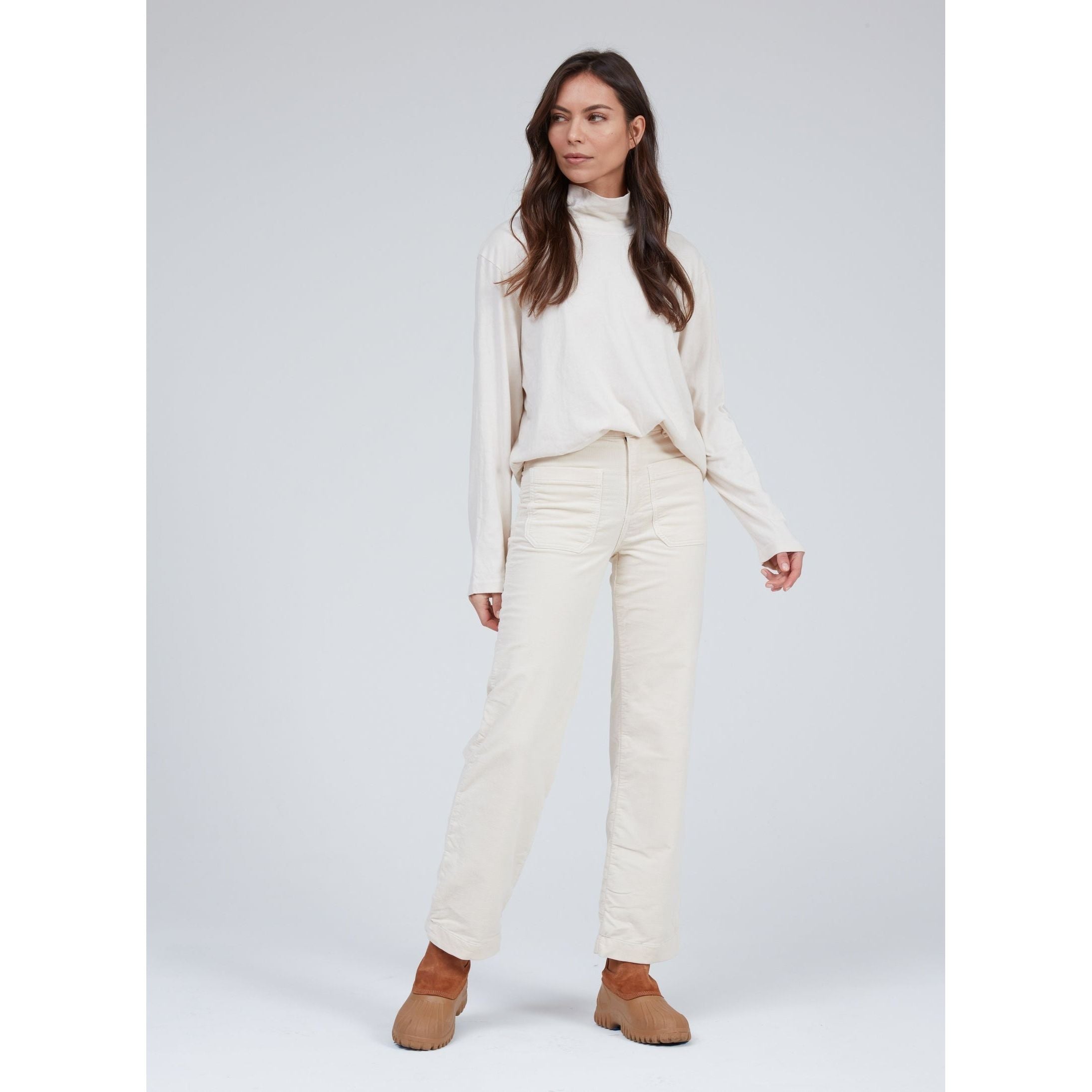 A person stands against a plain background wearing the ASKK NY Brighton Wide Leg Bone pants paired with a light-colored turtleneck and brown ankle boots. Made in Los Angeles, they have long, wavy hair and are looking to the side.