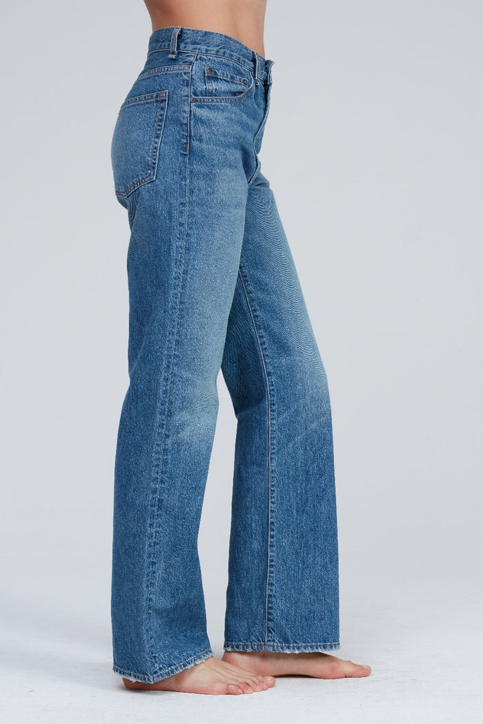 A person stands barefoot, wearing high-waisted, light blue ASKK NY Straight Jean Bush Baby made from premium Japanese denim. The jeans feature a classic five-pocket design with a subtle faded effect. The plain, light-colored background accentuates the side view of these stylish high-rise jeans from ASKK NY.