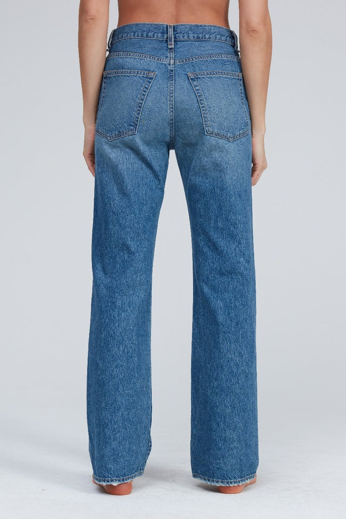 A person is standing facing away from the camera, wearing the ASKK NY Straight Jean Bush Baby. These high-waisted, straight-leg blue jeans are crafted from Japanese denim and feature back pockets with a central seam running down to the hem. The backdrop is plain and light-colored.