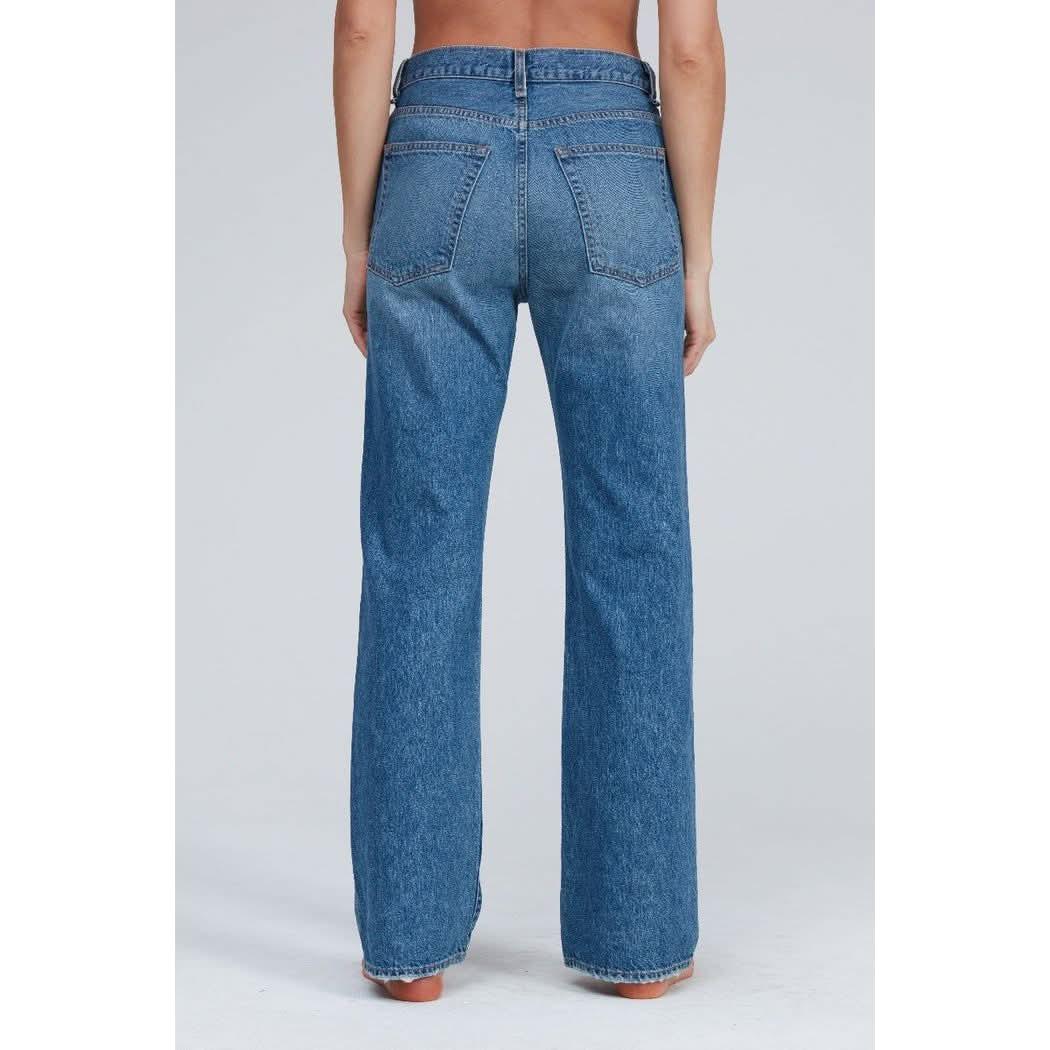 A person is standing facing away from the camera, wearing the ASKK NY Straight Jean Bush Baby. These high-waisted, straight-leg blue jeans are crafted from Japanese denim and feature back pockets with a central seam running down to the hem. The backdrop is plain and light-colored.