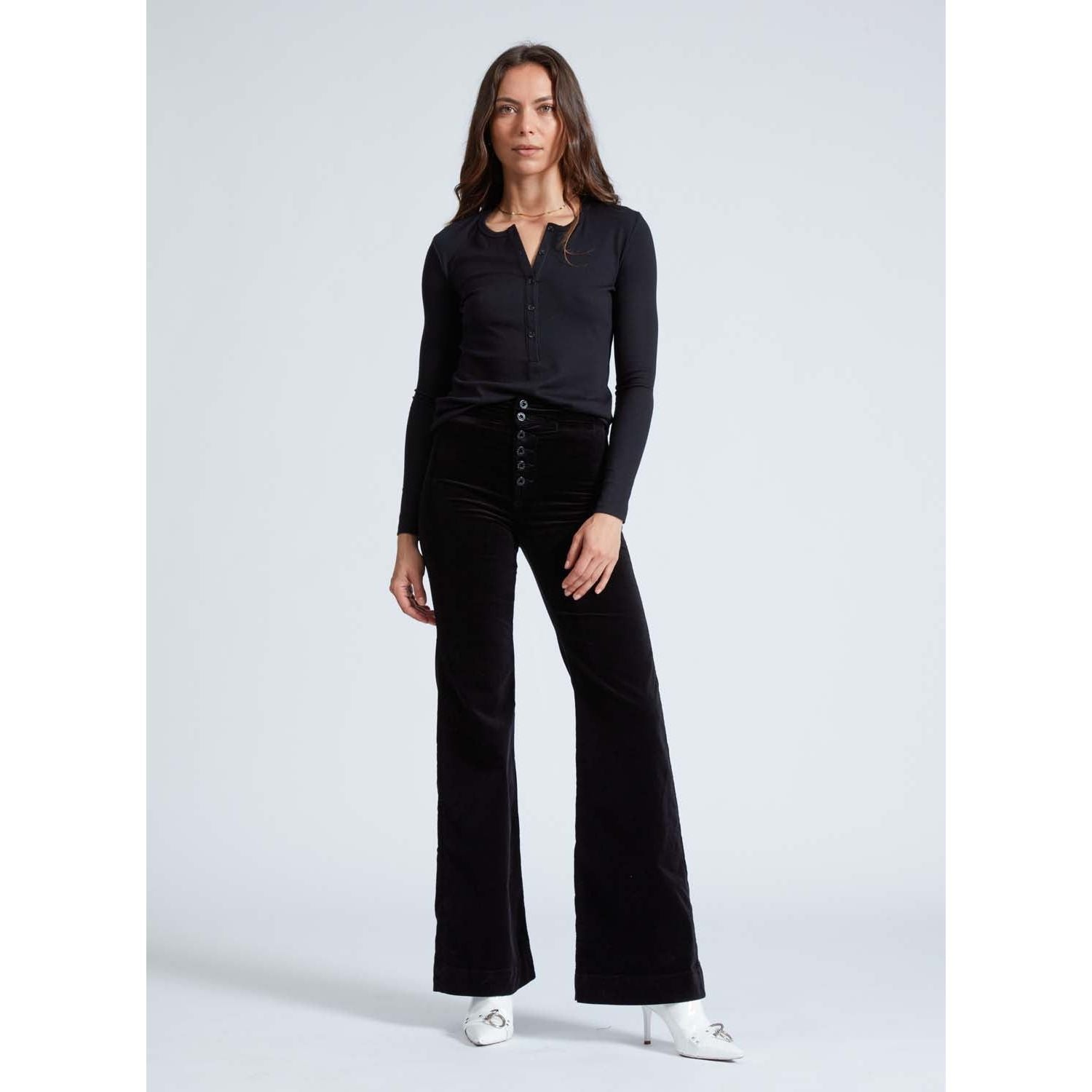 A woman with long hair stands against a plain background, showcasing the ASKK NY Brighton Wide Leg Black Velvet pants, paired with a black long-sleeve top. Her look is completed with crisp white shoes.