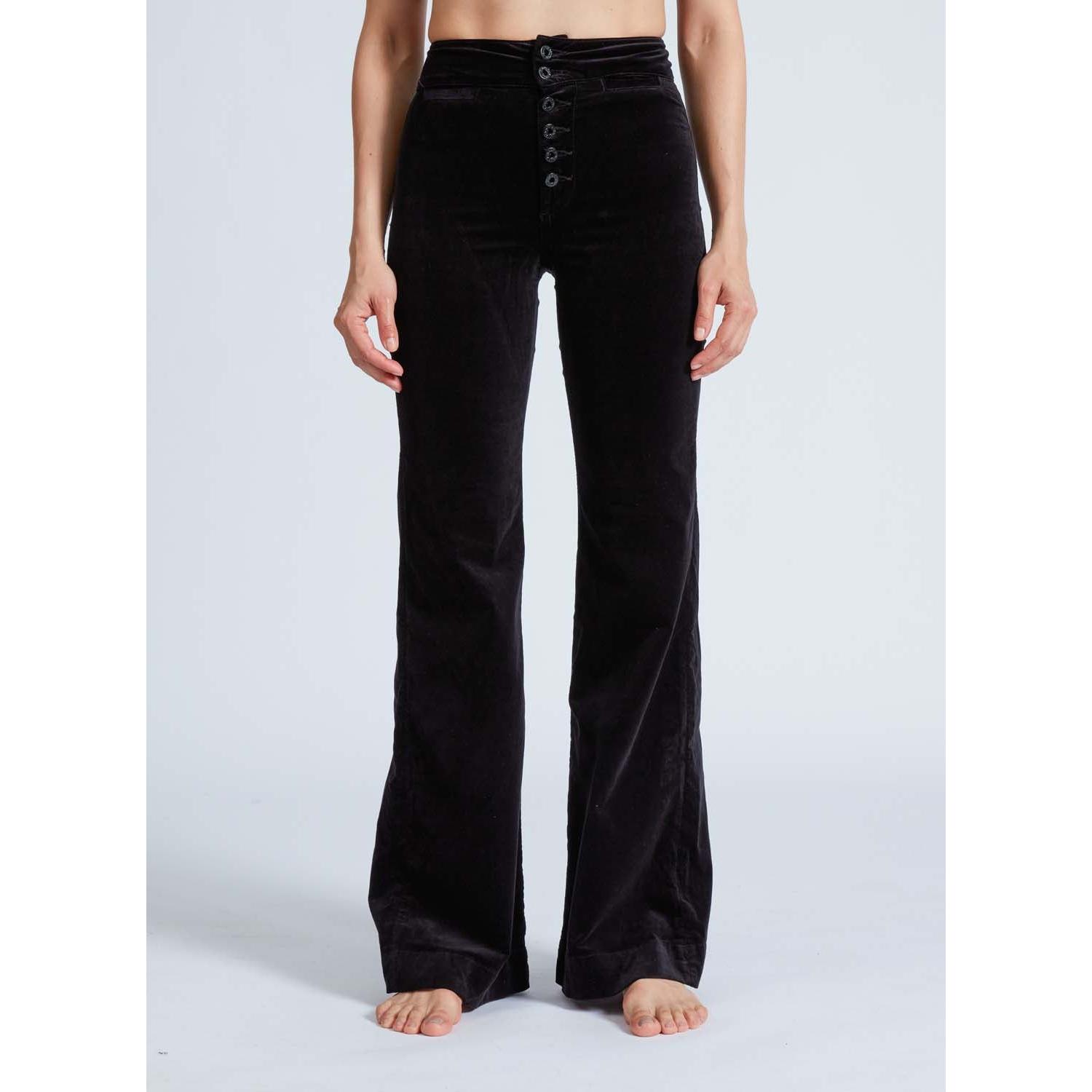 A person stands barefoot on a light background, wearing the ASKK NY Brighton Wide Leg Black Velvet pants. These high-waisted pants feature a button-up front and a sleek finish. The person's hands rest relaxed by their sides.