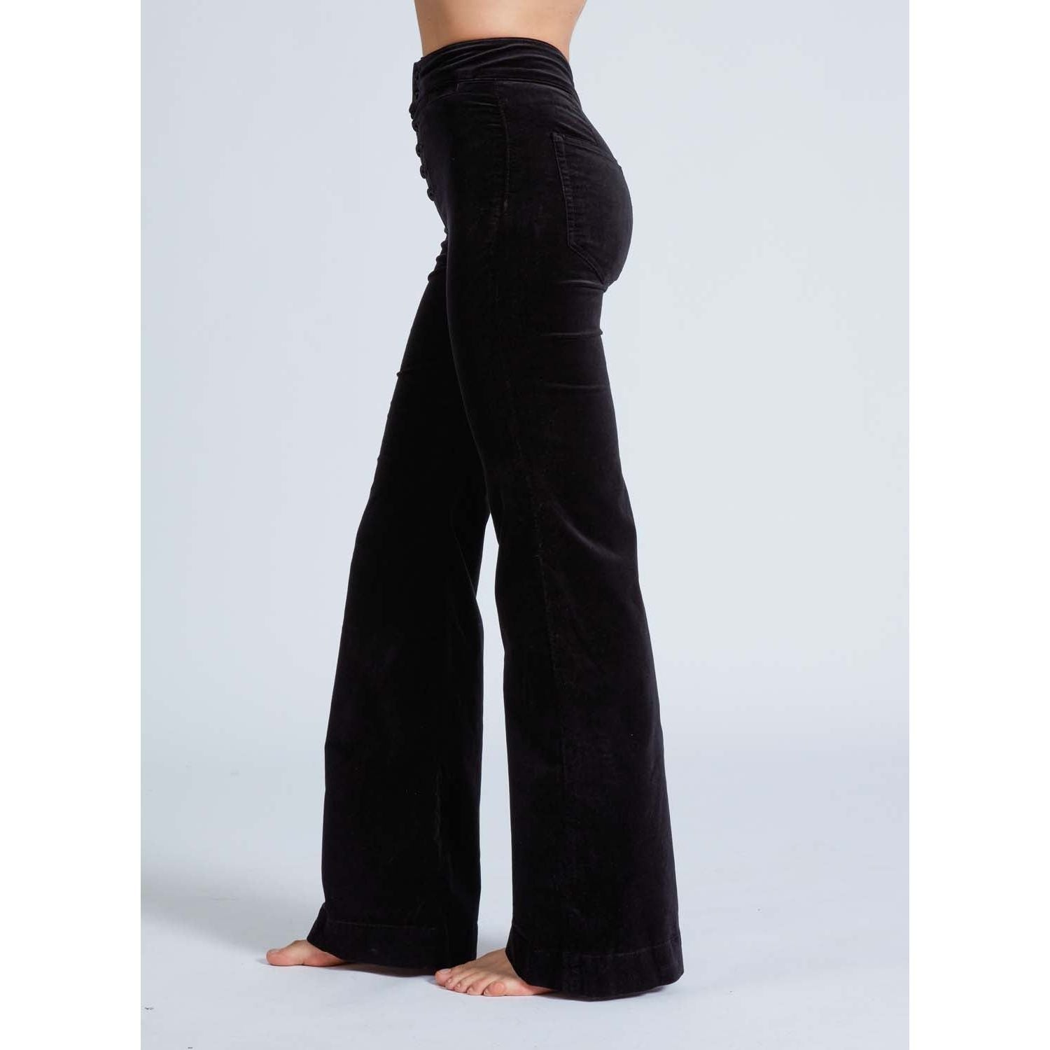 A person stands barefoot on a light background, wearing the ASKK NY Brighton Wide Leg Black Velvet pants. These high-waisted pants have a wide flare and smooth texture, creating a casual yet stylish appearance.