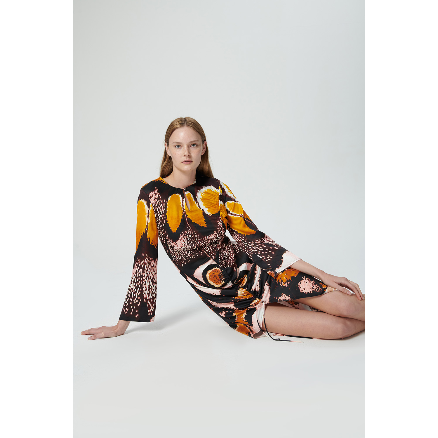 A woman with long blonde hair is seated on the floor against a plain background. She is wearing the Chufy Orchid Maxi Dress, which showcases a vibrant pattern of orange, yellow, white, and black. Made from sustainable fabric by Chufy, this dress features long sleeves and a draped design. She gazes directly at the camera with a calm expression.