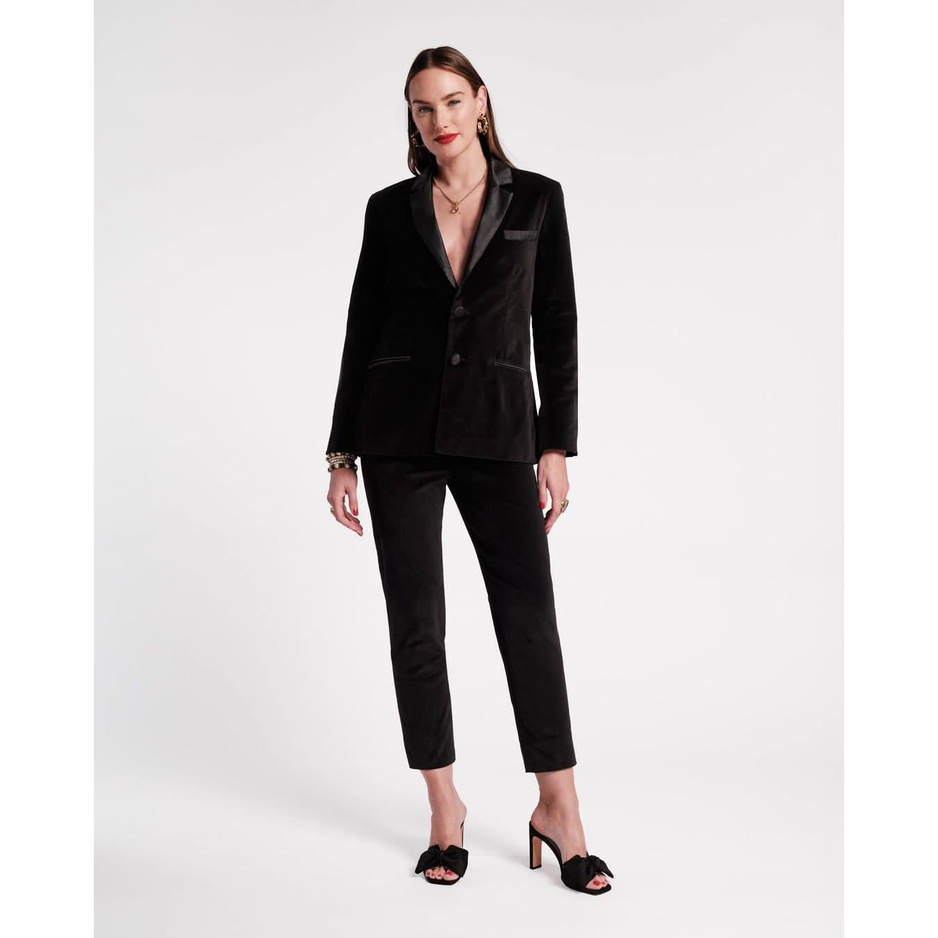 A person stands against a plain background, showcasing the Frances Valentine Tuxedo Jacket in black, featuring a sleek velvet design with a deep neckline. Complementing the look with matching high heels, they have long hair and are adorned with gold bracelets and earrings, presenting a tailored silhouette that exudes elegance.