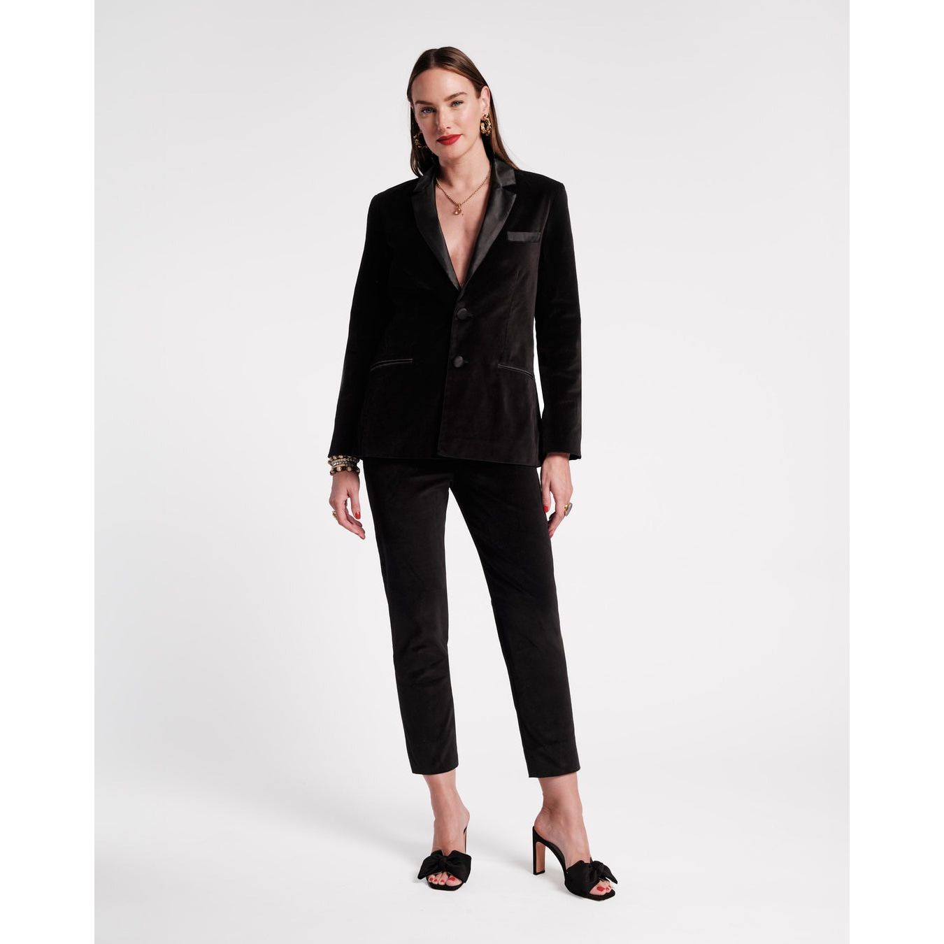 A person stands confidently against a plain background, dressed in the elegant Frances Valentine Lucy Pant Velvet suit featuring a satin lapel. They have long brown hair and are accessorized with gold bracelets and black high-heeled shoes.