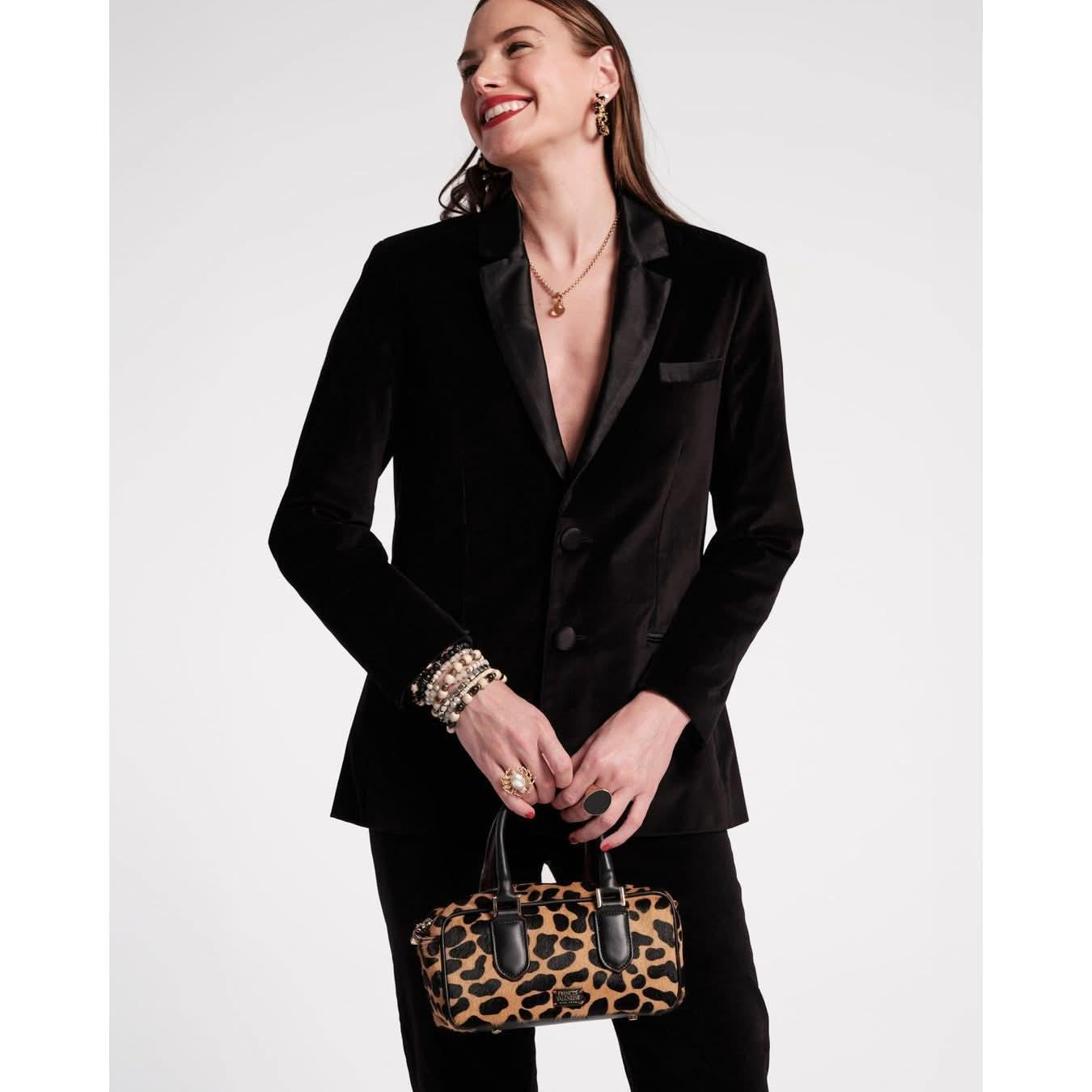 A woman in a Frances Valentine Tuxedo Jacket crafted from stretch velvet in sleek black smiles while holding a leopard print handbag. She accessorizes with gold jewelry, including earrings, a necklace, and bracelets. Her long hair cascades over her shoulders as she poses against a plain white background.