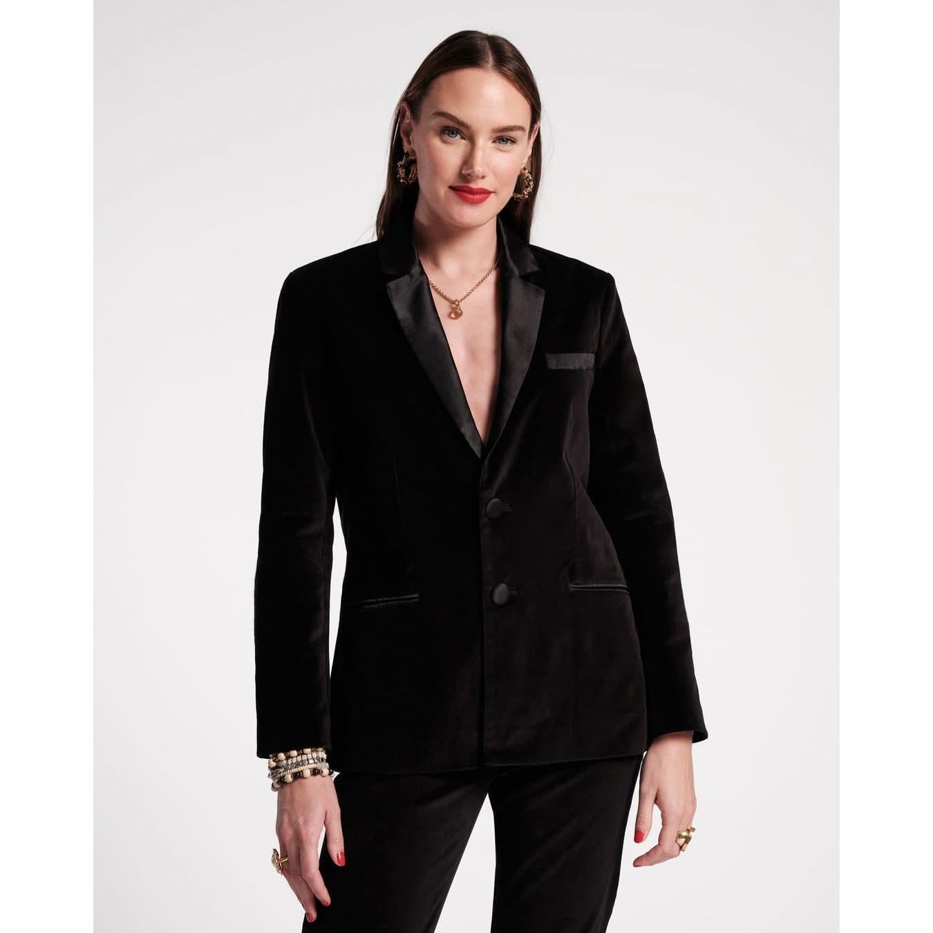 A person with long hair and red lipstick is wearing a Frances Valentine Tuxedo Jacket made from luxurious stretch velvet in black, paired with matching pants. They are accessorized with bracelets, earrings, and a necklace, striking a pose against a plain white background.