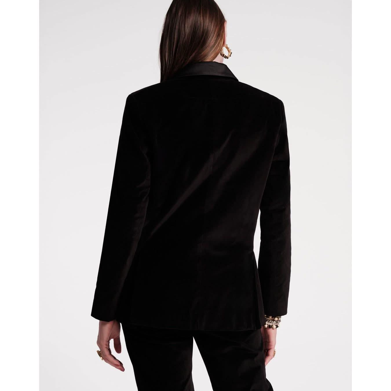 A person with long brown hair is seen from the back wearing the Frances Valentine Tuxedo Jacket in black, characterized by its stylish satin lapel and smooth texture. They accessorize elegantly with gold hoop earrings and a few gold bracelets on their wrist.